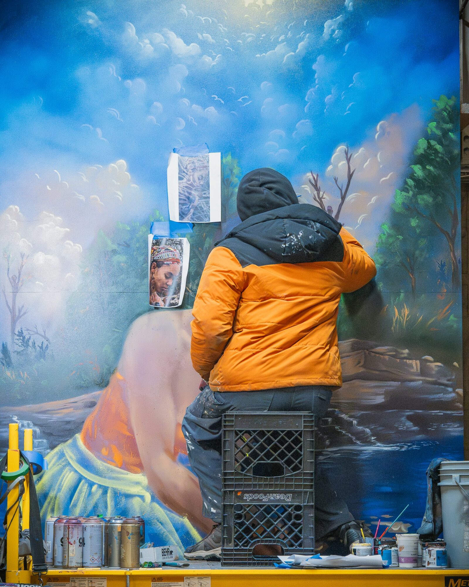 Person in orange jacket and black hood facing a nature scene with trees and water, with art supplies nearby.