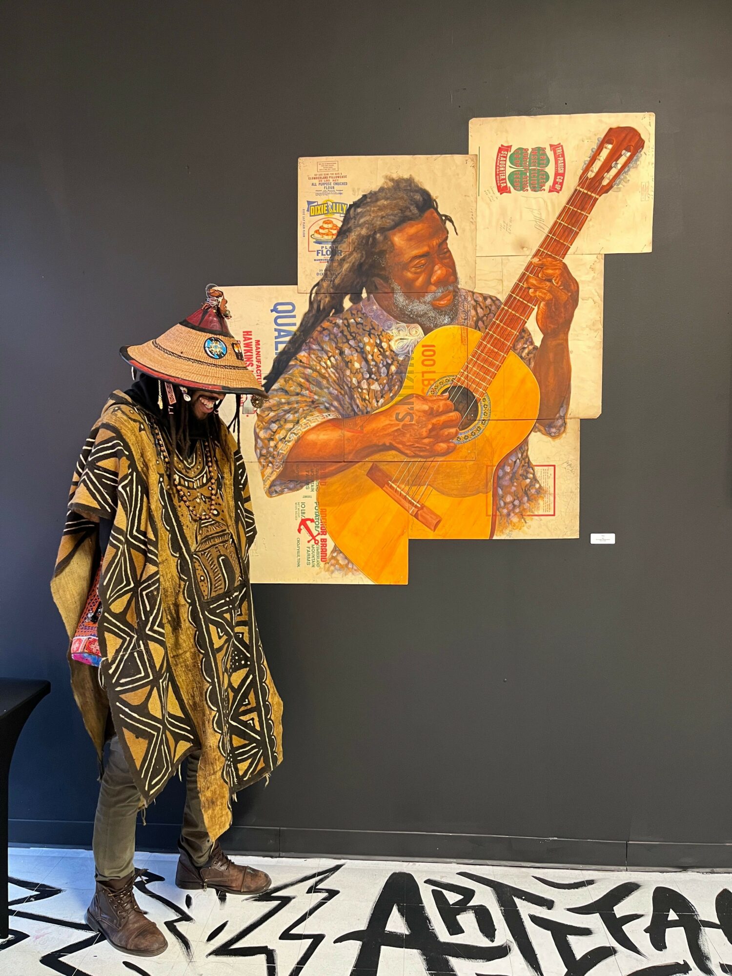 Person with a painted face playing guitar, wearing patterned clothing, standing against a dark background.