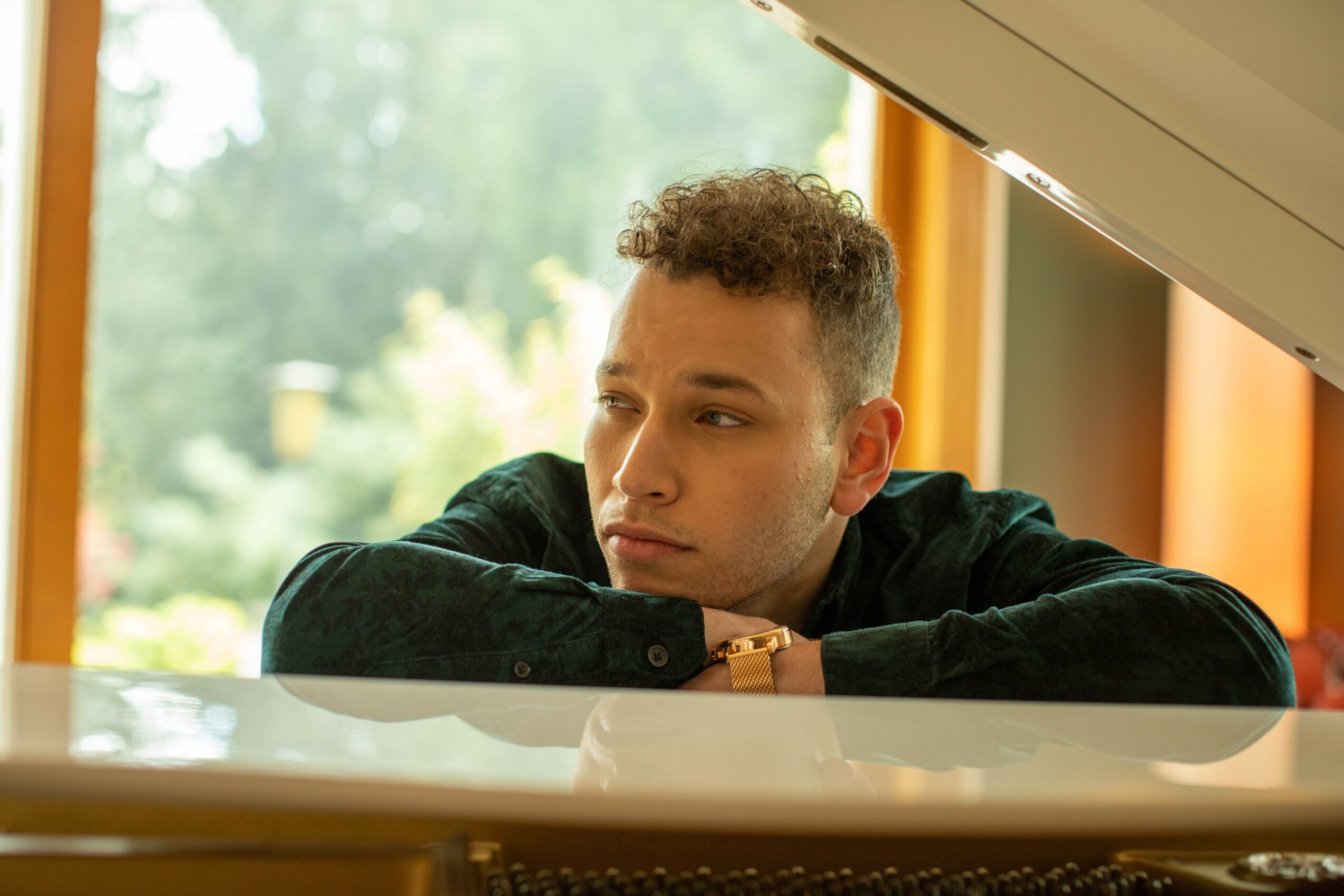 Young man with curly hair resting his arms on a white piano, looking out a window with trees outside.