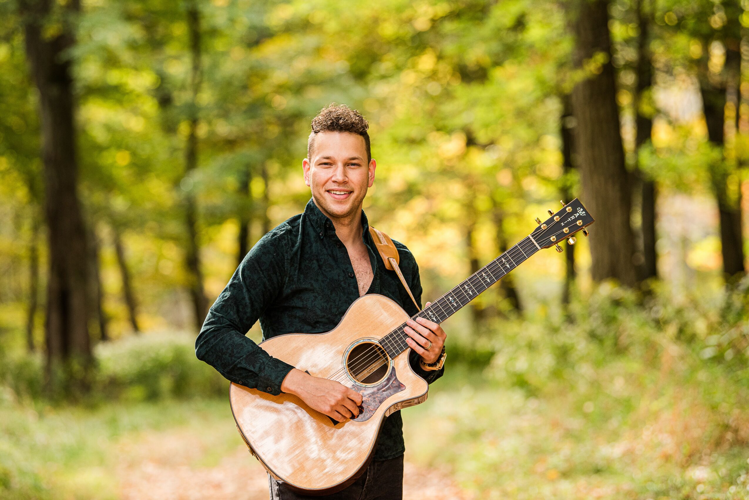 Man holding an acoustic guitar outdoors in a forest with green and yellow leaves