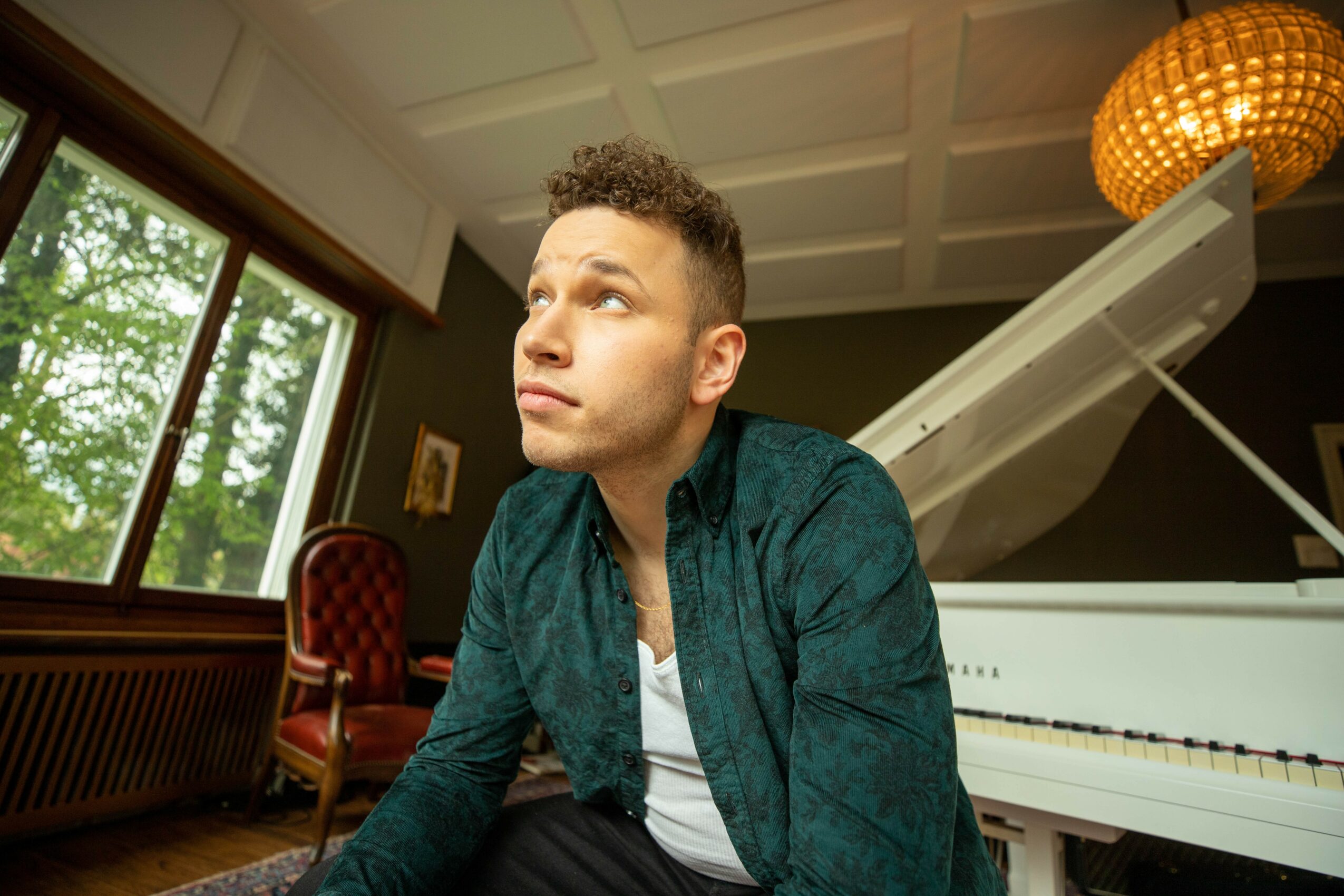Young man with short curly hair looking out a window in a room with a white grand piano and a red chair.