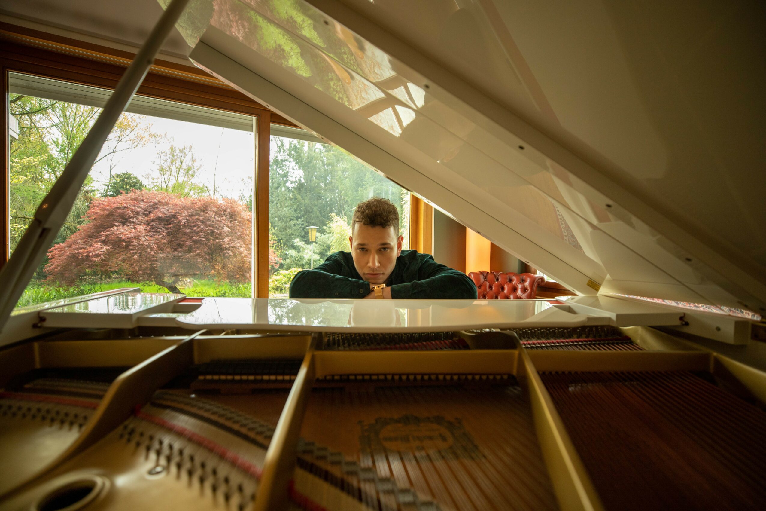 Person sitting at a white grand piano with open lid, looking at the camera, in a room with large windows and greenery outside.