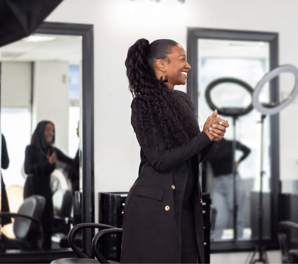 Smiling woman with curly hair in a black coat, clapping in a room with mirrors and chairs.