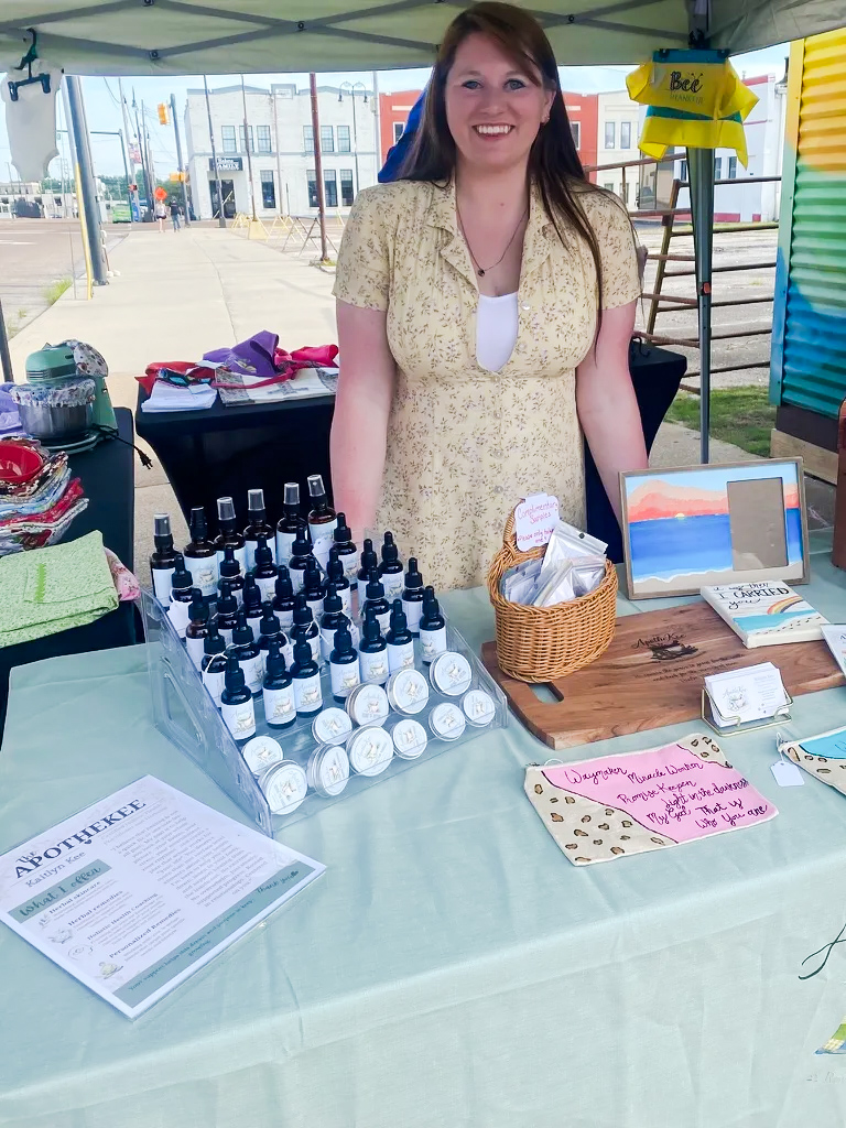 Smiling woman standing behind a table with small bottles, a basket, and signs at an outdoor market stall.