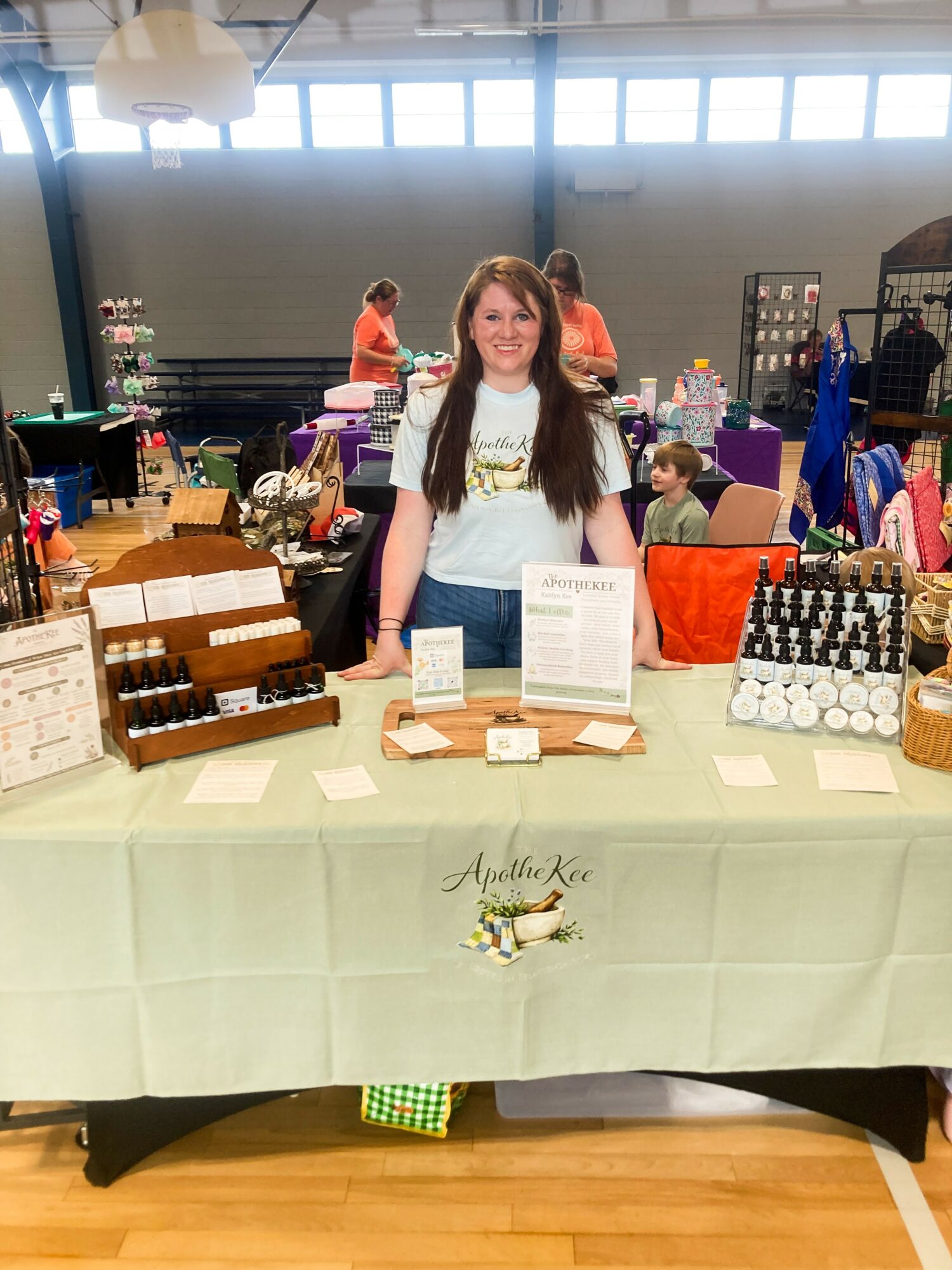Young woman standing behind a table with various items at an indoor market or fair, with people and booths in the background.