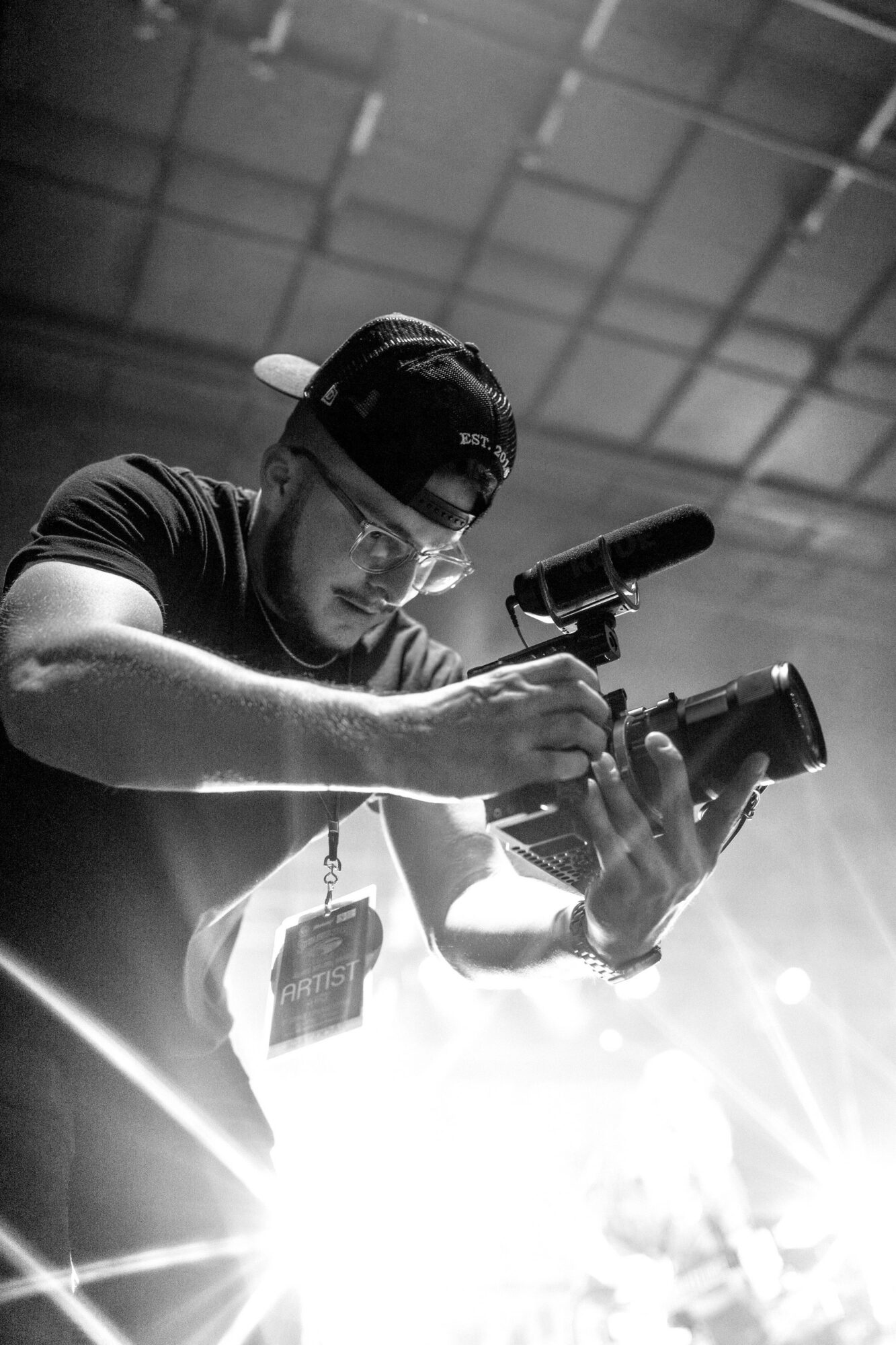 Person wearing glasses and a cap, holding a camera and a microphone, indoors with a ceiling grid in background.