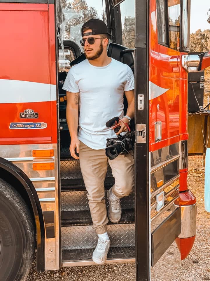 Man with sunglasses, beard, white t-shirt, beige pants, white sneakers, holding a camera, standing on bus steps outside.