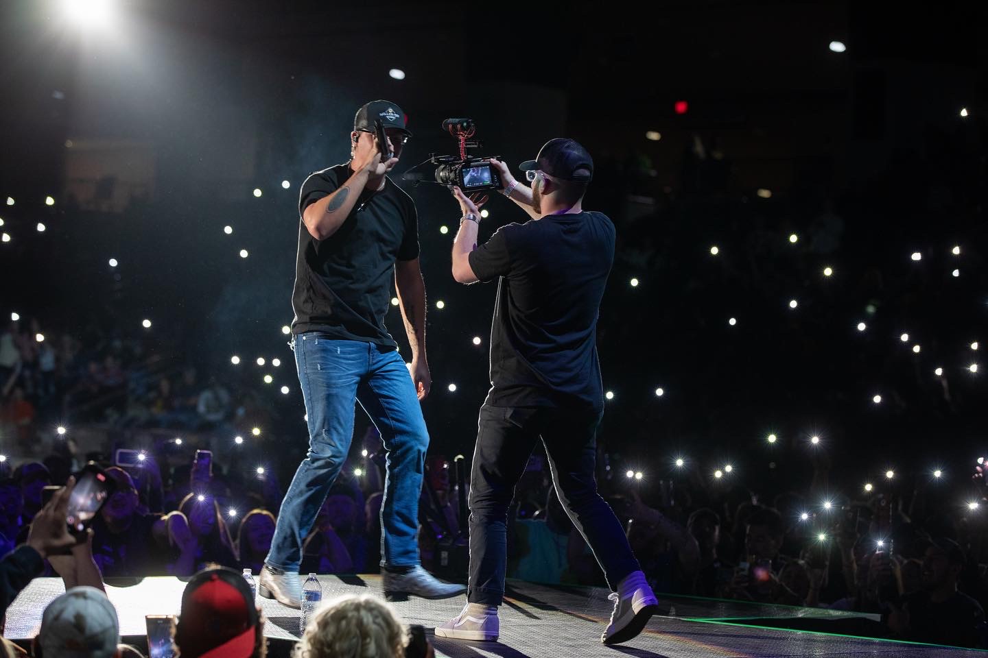 Two performers on stage, one holding a microphone and the other filming, with an audience using lights and phones in the background.