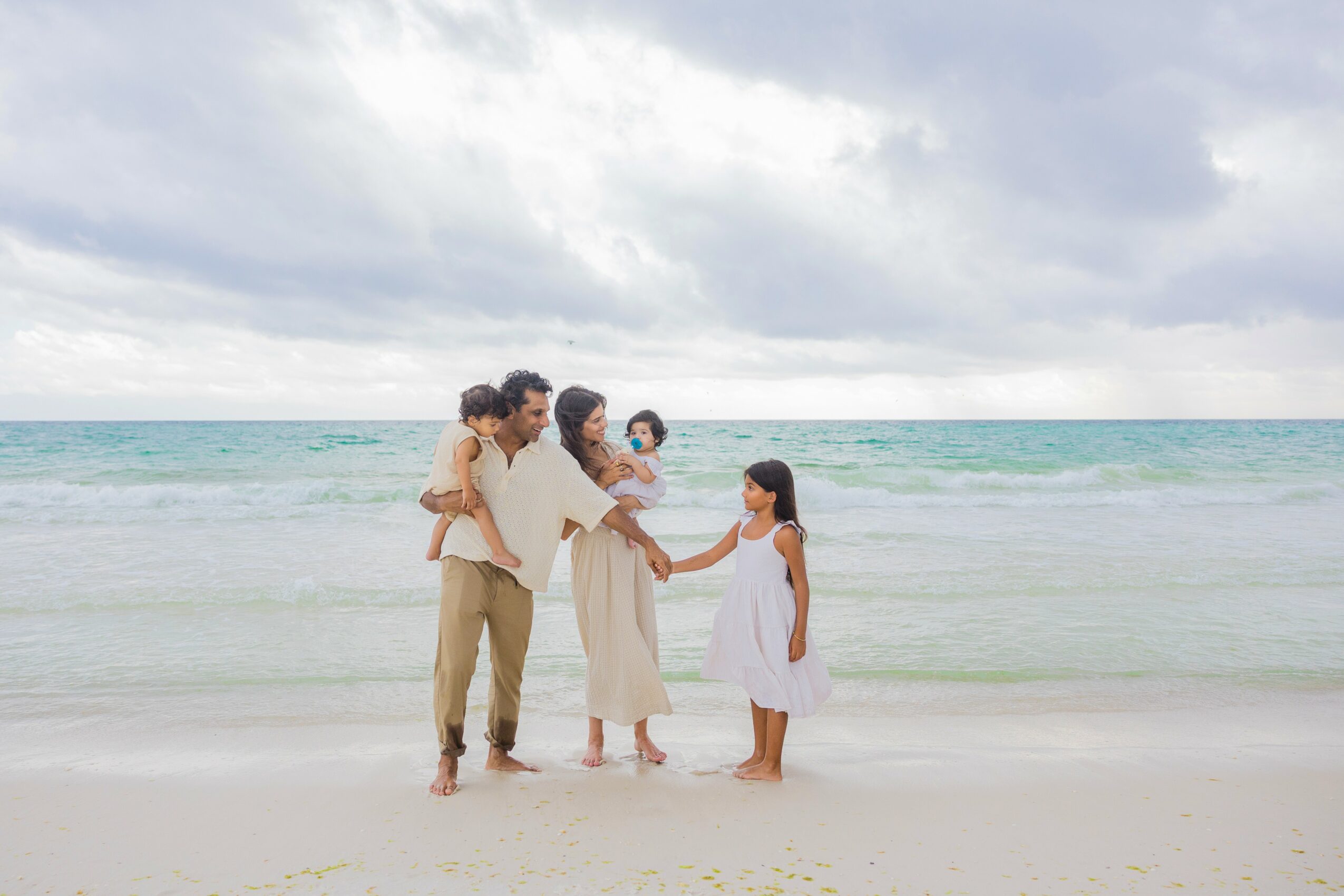 Group of five people, including children, standing on a beach near the ocean, holding hands and interacting under cloudy sky.
