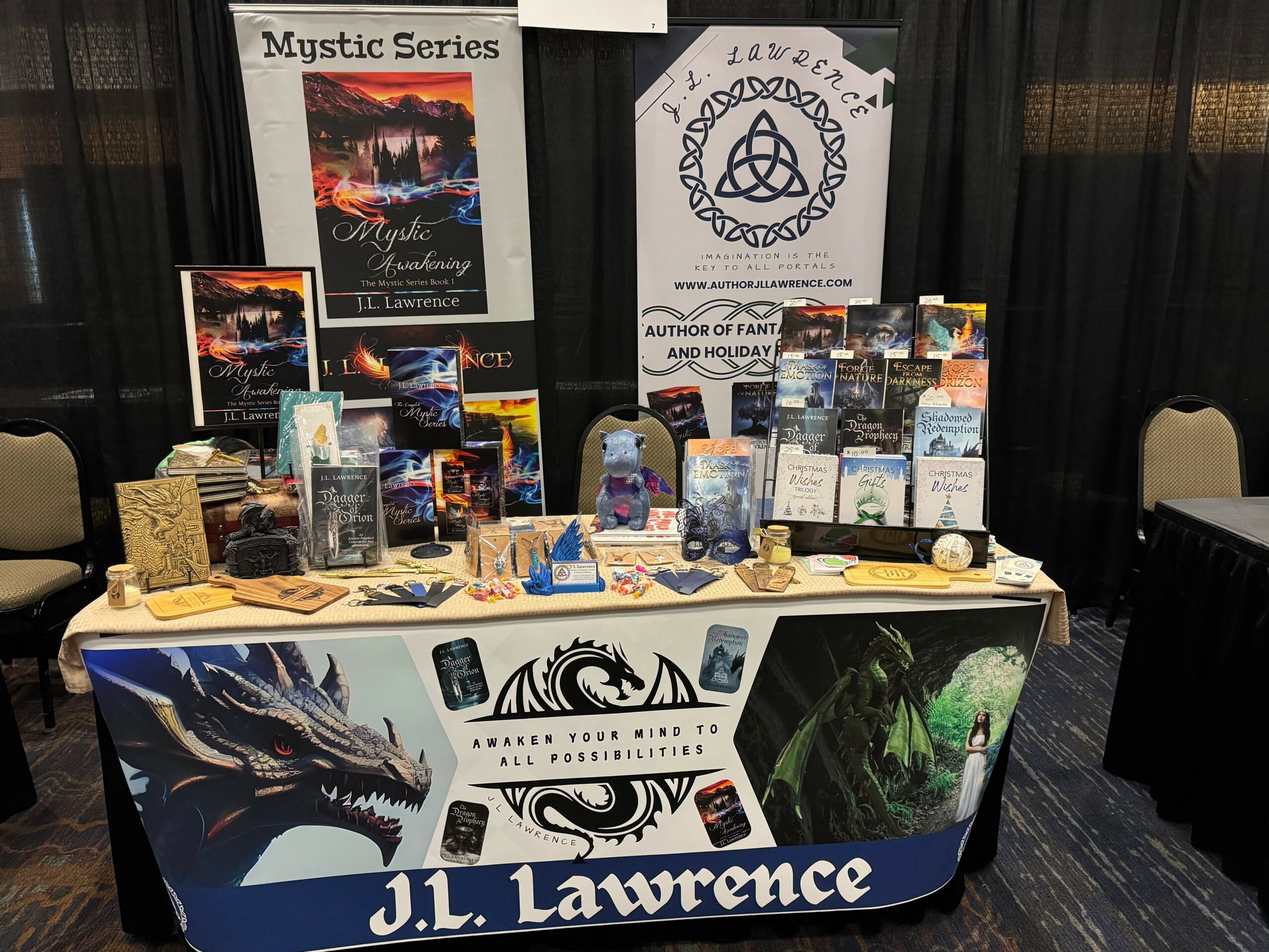 Book display table with books, posters, and banners, including Mystic Series and author J.L. Lawrence, set against black curtains.