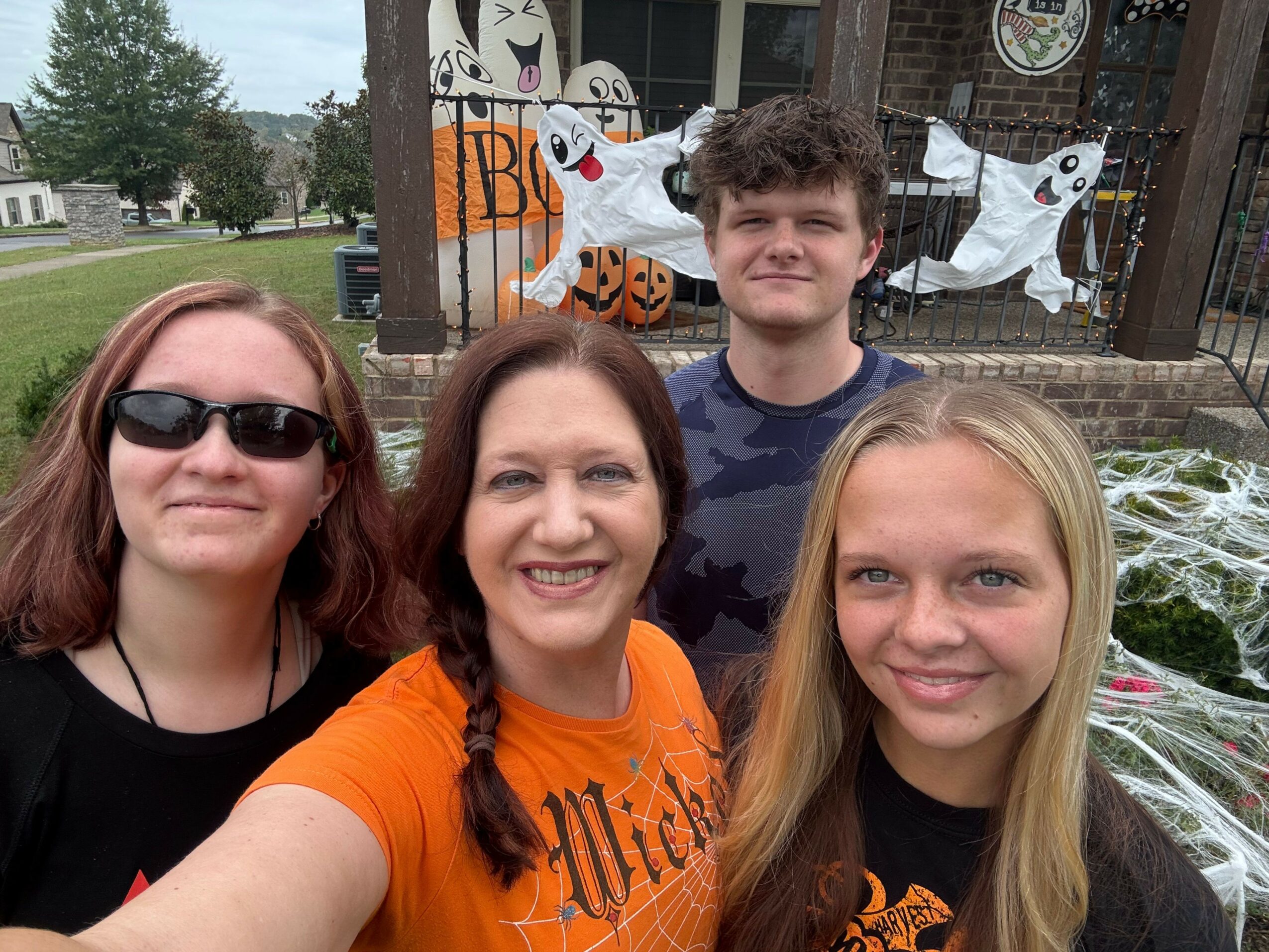 Four women smiling outdoors, standing in front of a white vehicle with graffiti, trees, and buildings in the background.