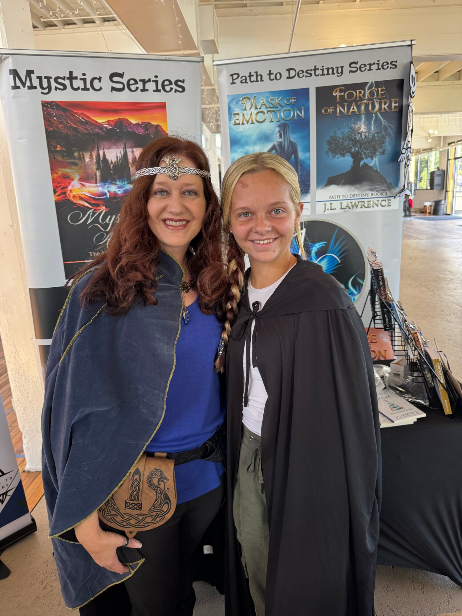 Two women smiling, standing in front of book displays, one in a blue dress and cape, the other in a graduation gown.