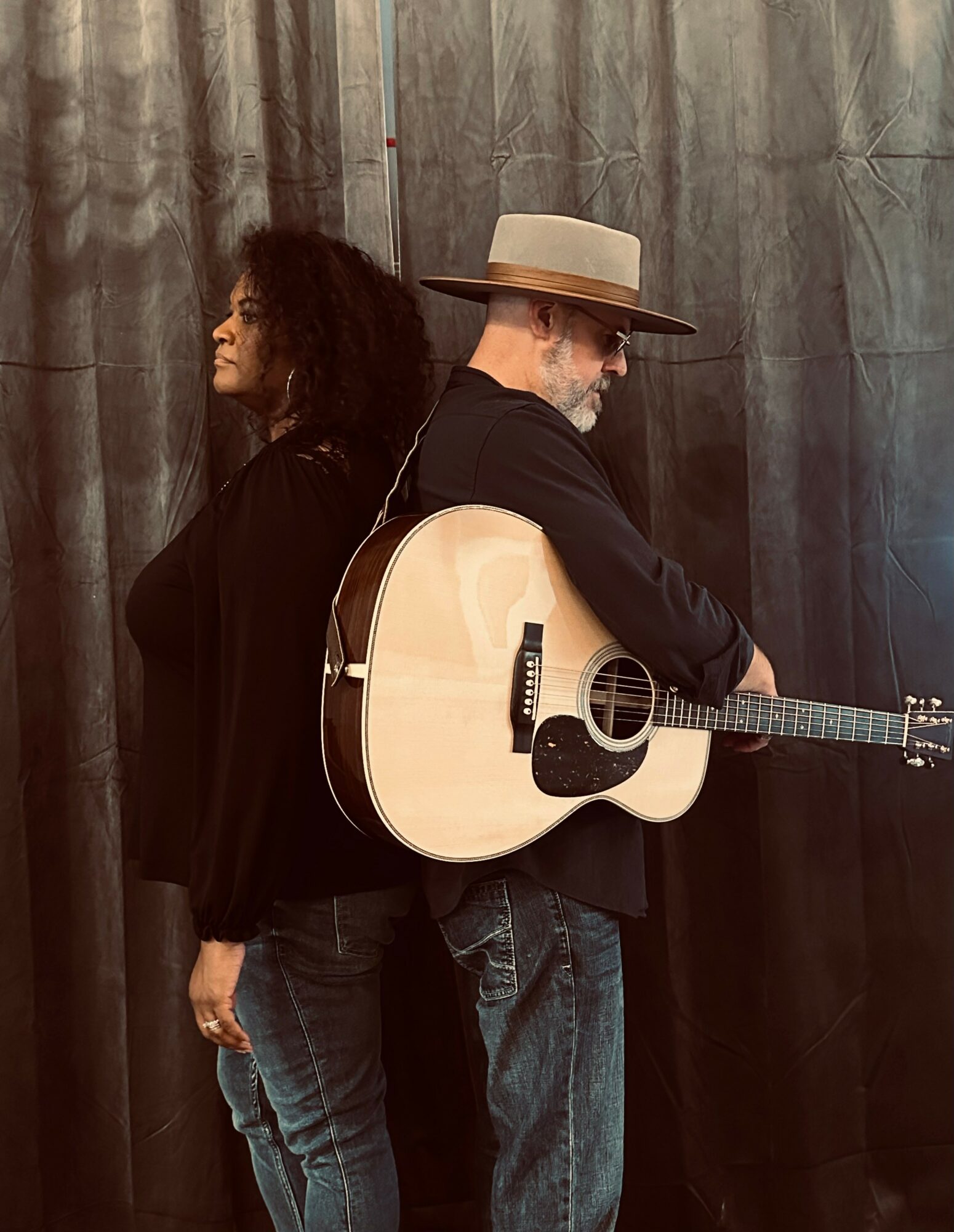 Two people back-to-back, one with curly hair and the other with a hat and guitar, standing in front of a curtain.