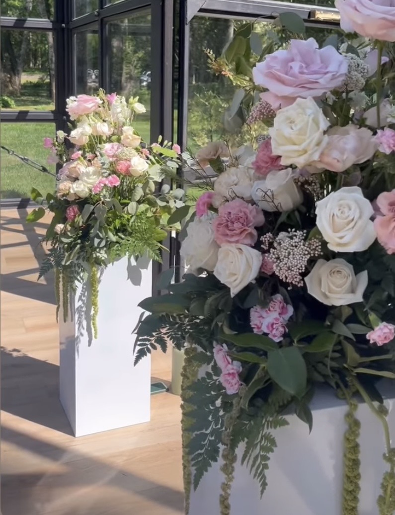 Two large floral arrangements with pink and white flowers in white vases inside a glass-enclosed space.