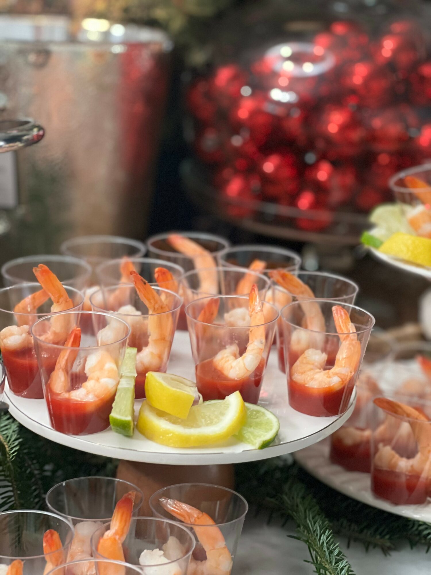 Shrimp cocktails with lemon wedges and sauce in small cups on a serving tray, with a blurred background of red berries.