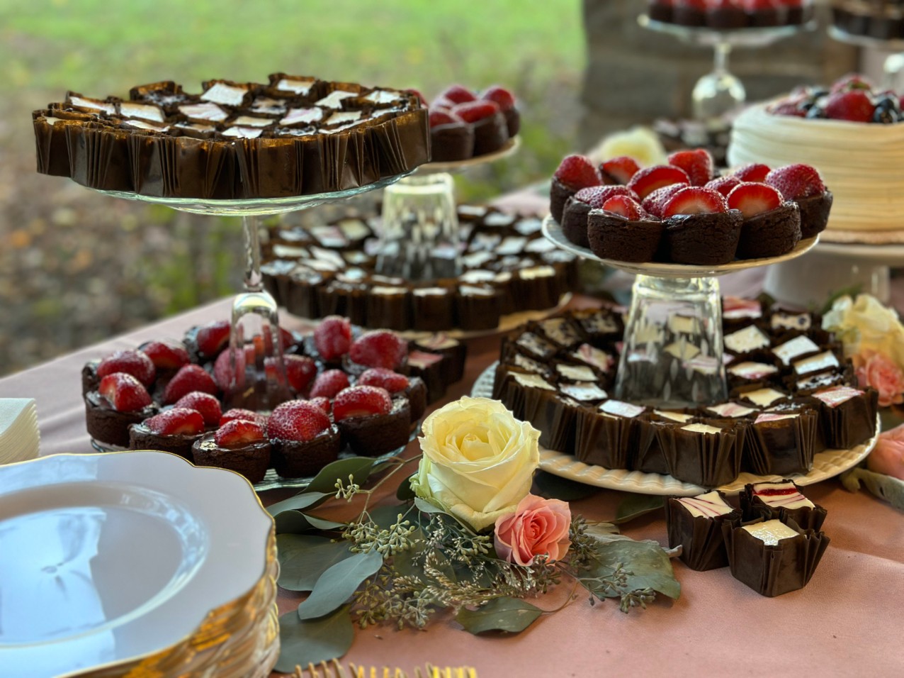 Dessert table with cakes, chocolates, and berries, decorated with a white and pink rose, outdoors.