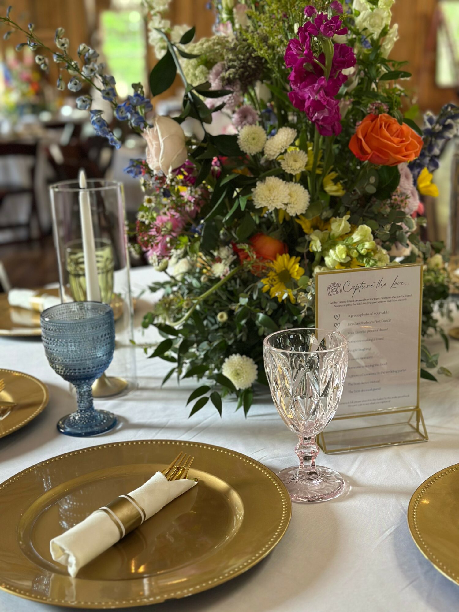 Table setting with a large floral centerpiece, gold plates, a blue glass, and a pink glass, with a menu card.