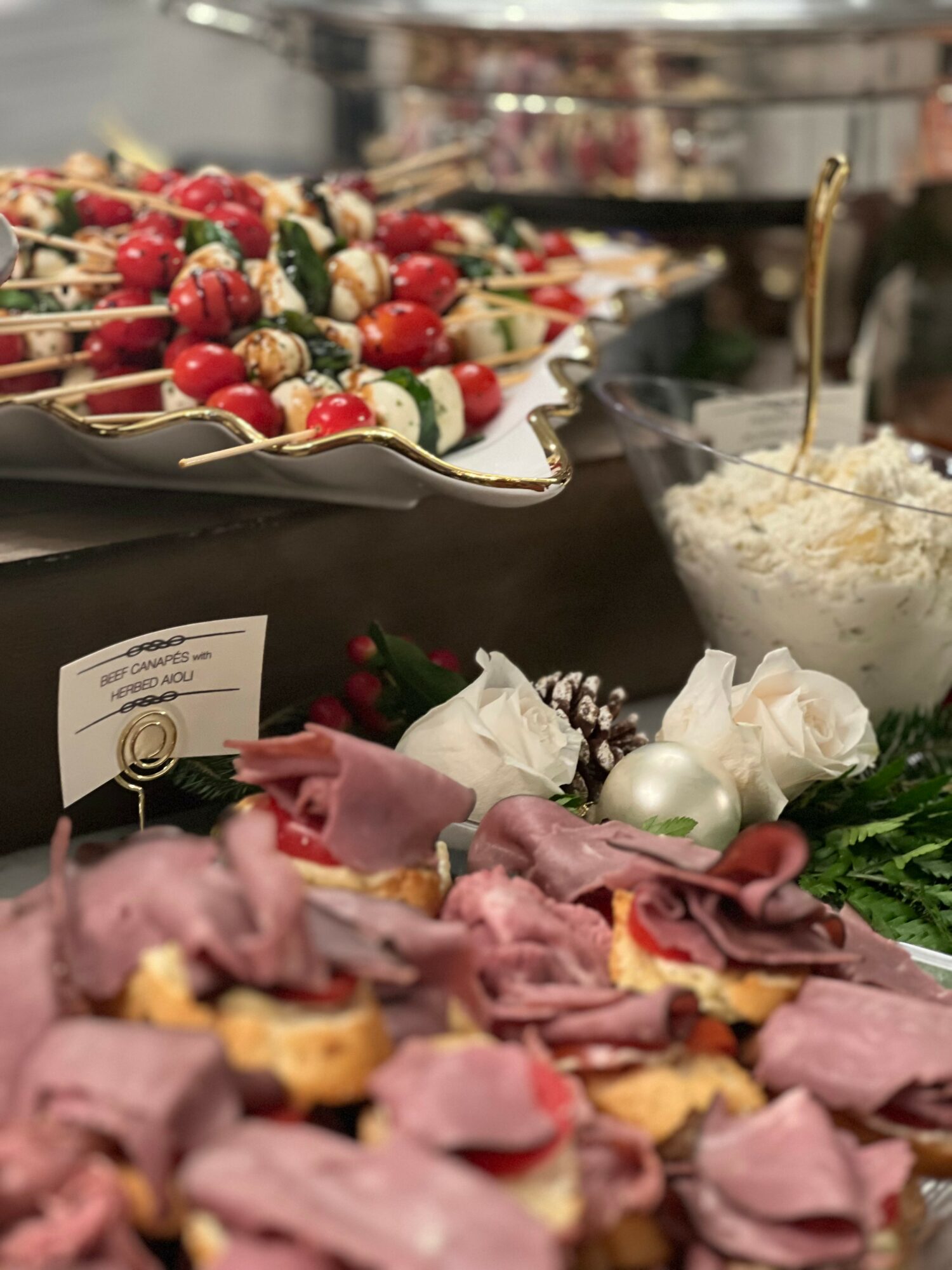 Skewers with cherry tomatoes, mozzarella, and basil, a bowl of rice salad, and sliced ham with garnishes on a table.