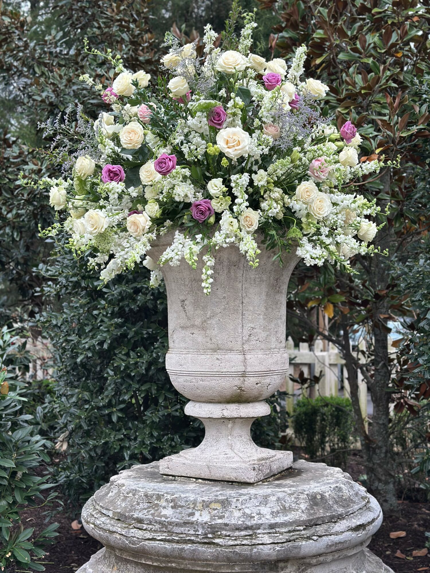 Large stone urn filled with white and purple flowers, placed outdoors with trees and greenery in background.