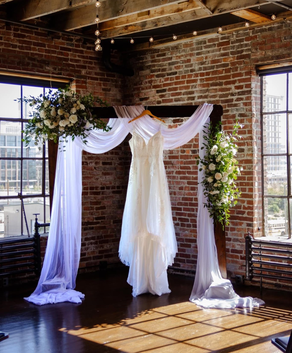 Wedding dress hanging between two floral arrangements with white fabric drapes, brick wall background, sunlight streaming through windows.
