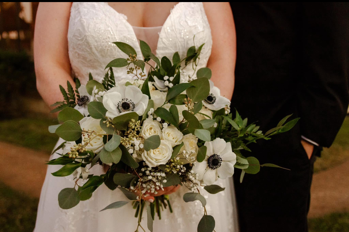 Person in a white dress holding a large bouquet of white flowers and greenery, with a person in dark clothing nearby.