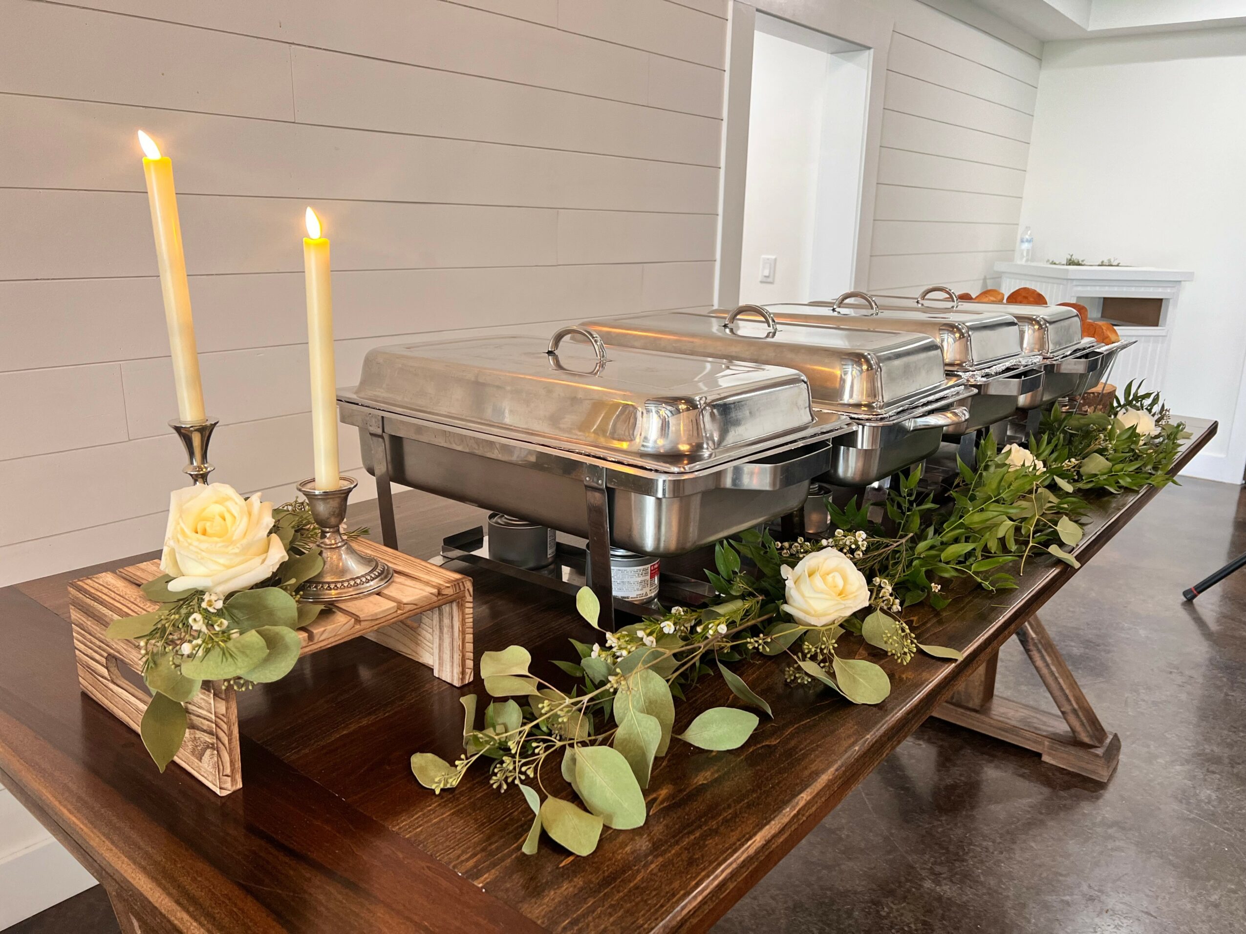 Buffet table with chafing dishes, candles, and floral decorations in a bright room.