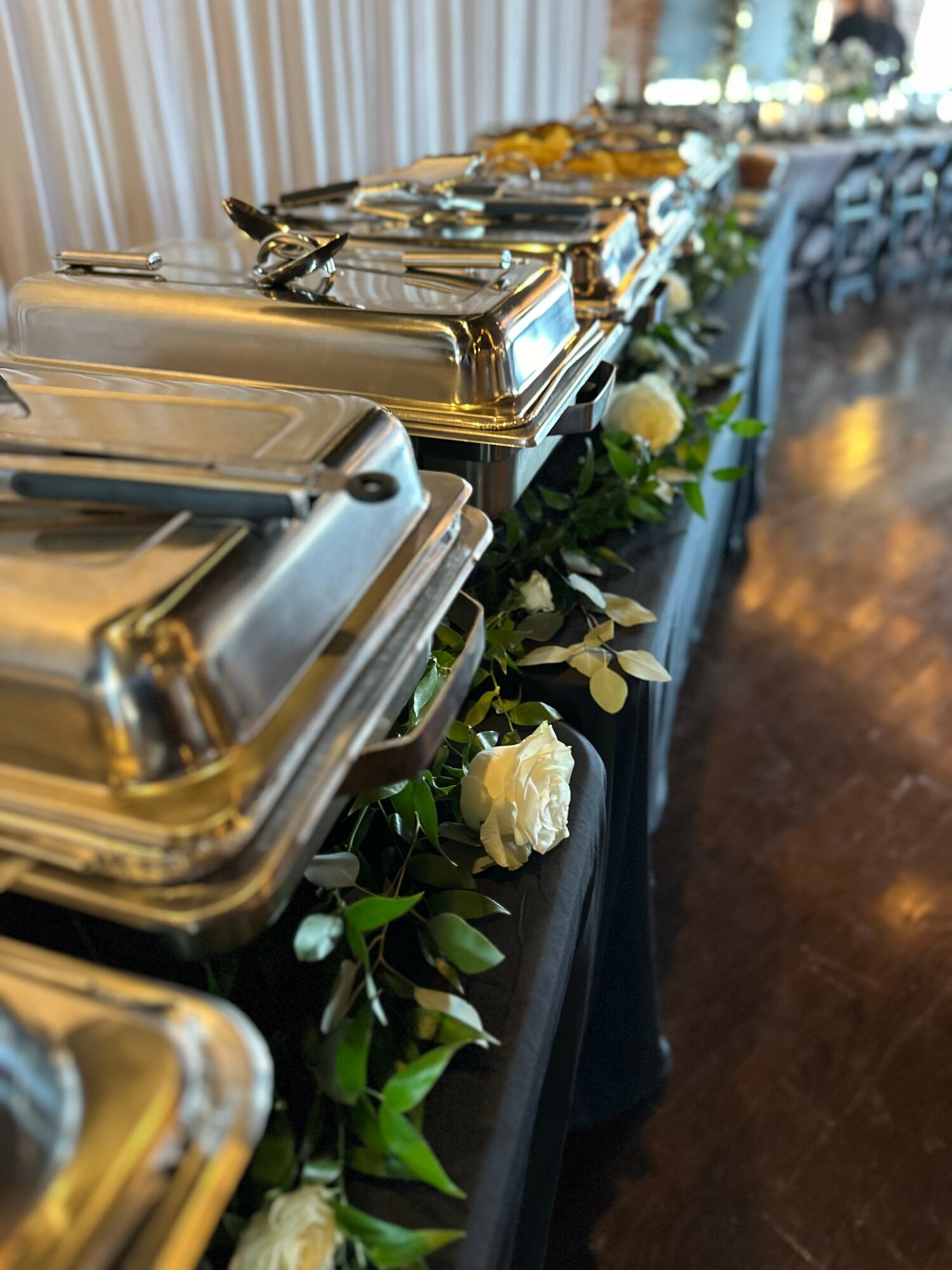 Buffet table with chafing dishes and floral decorations, set in a well-lit room with wooden flooring.