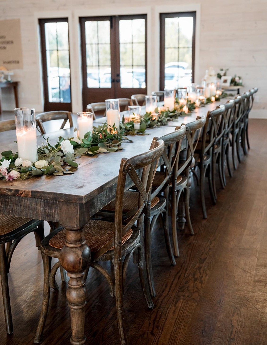 Long wooden table decorated with candles, flowers, and greenery, set in a bright room with large windows and wooden floor.
