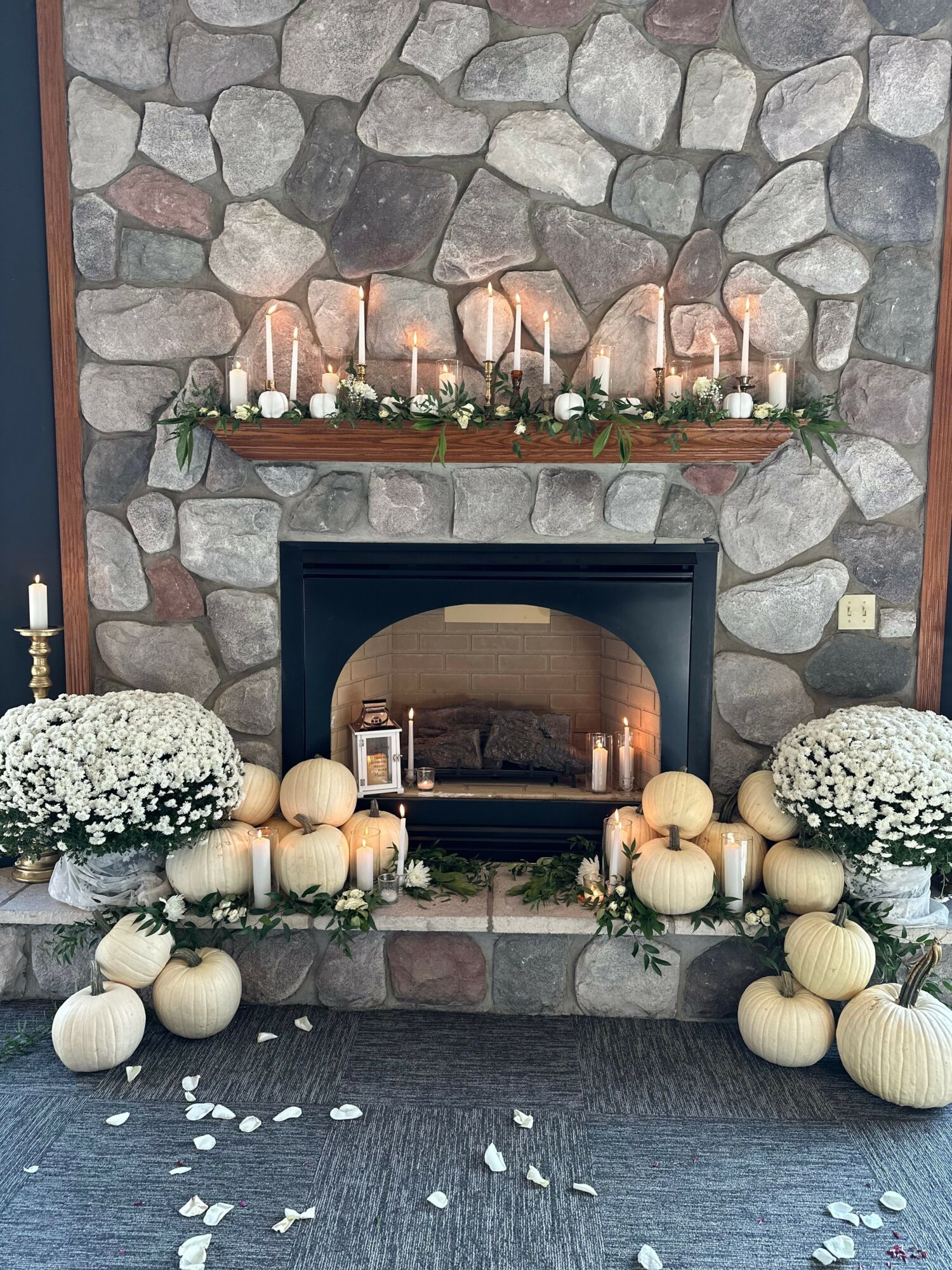 Decorative fireplace with candles, pumpkins, and flowers on a stone wall, with scattered white petals on the floor.