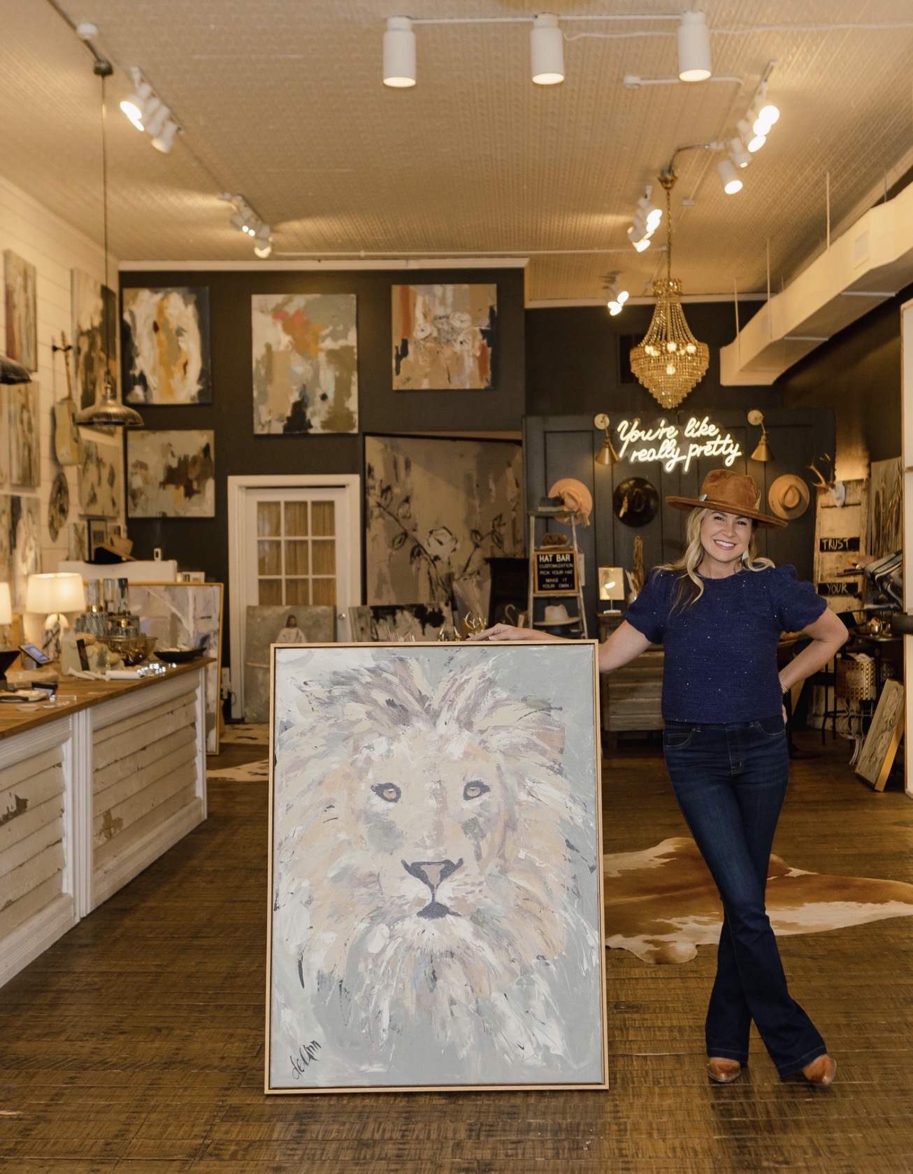 Woman standing in art gallery next to a large lion painting on an easel, smiling, with artwork displayed on walls behind her.