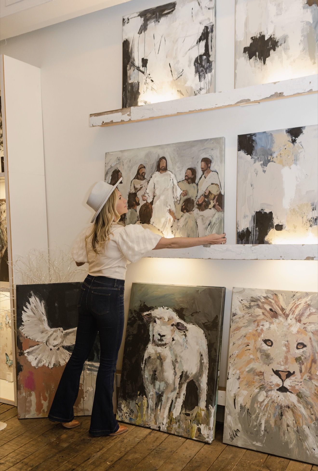 Woman arranging paintings on wall in art gallery, with animal and abstract artwork displayed.
