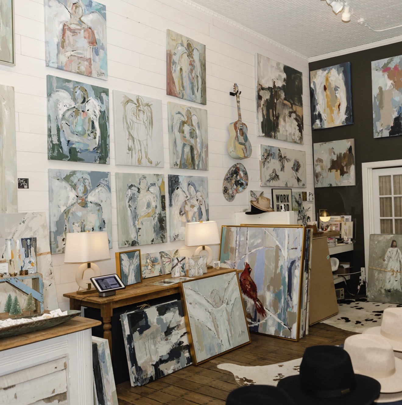 Gallery wall with abstract paintings, a guitar, and a table with lamps and framed artwork, in a cozy room.