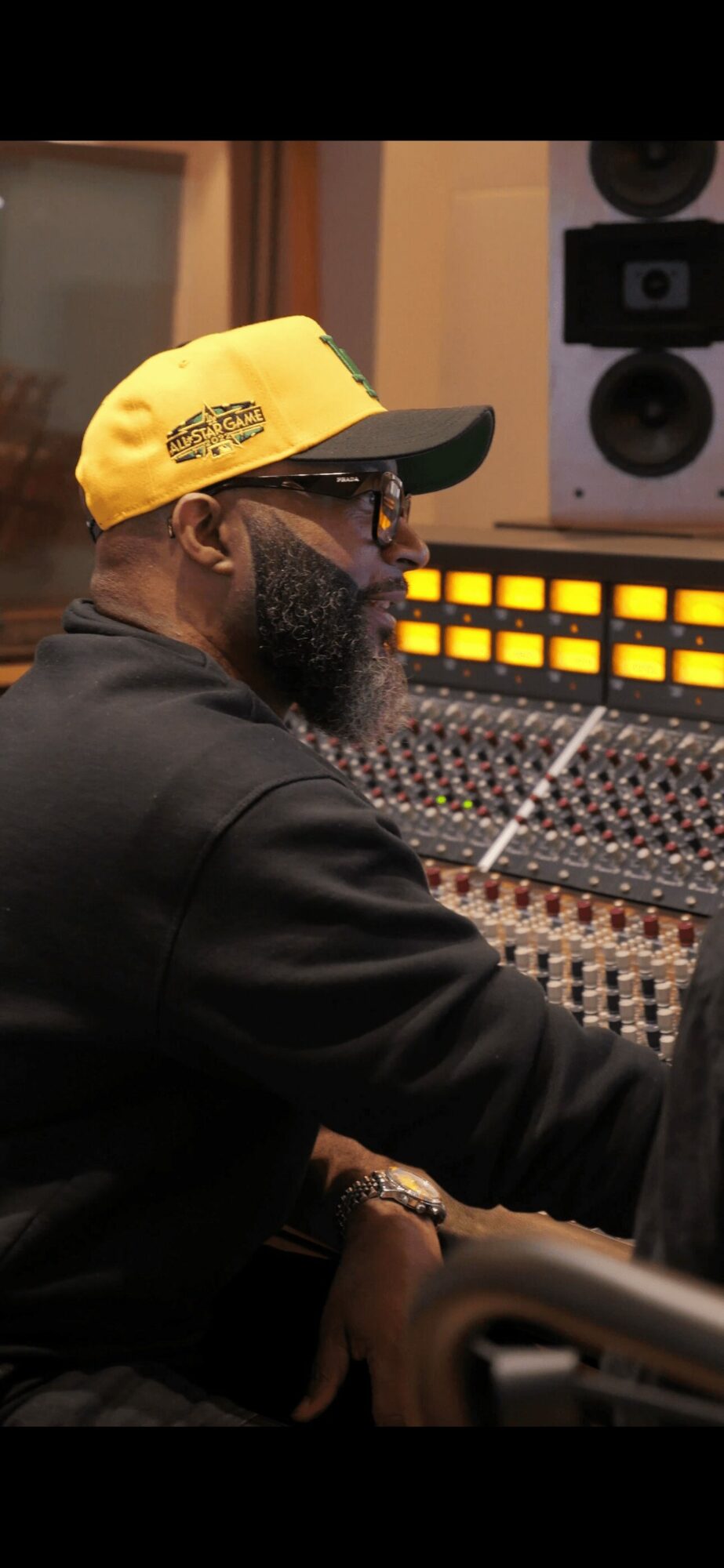 Man with a beard wearing a yellow cap and glasses working at a sound mixing console in a studio.