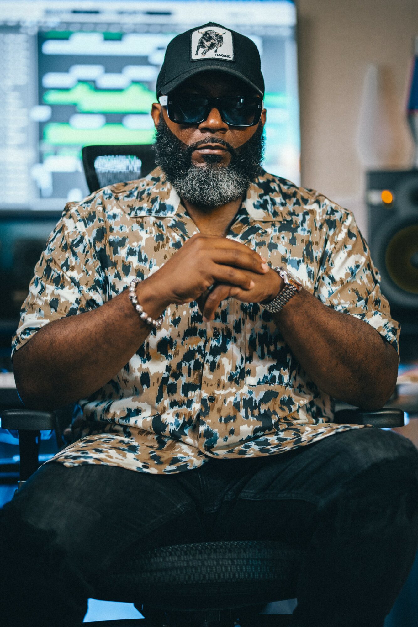 Man with sunglasses, beard, wearing a patterned shirt and cap, sitting in a studio with music equipment behind him.