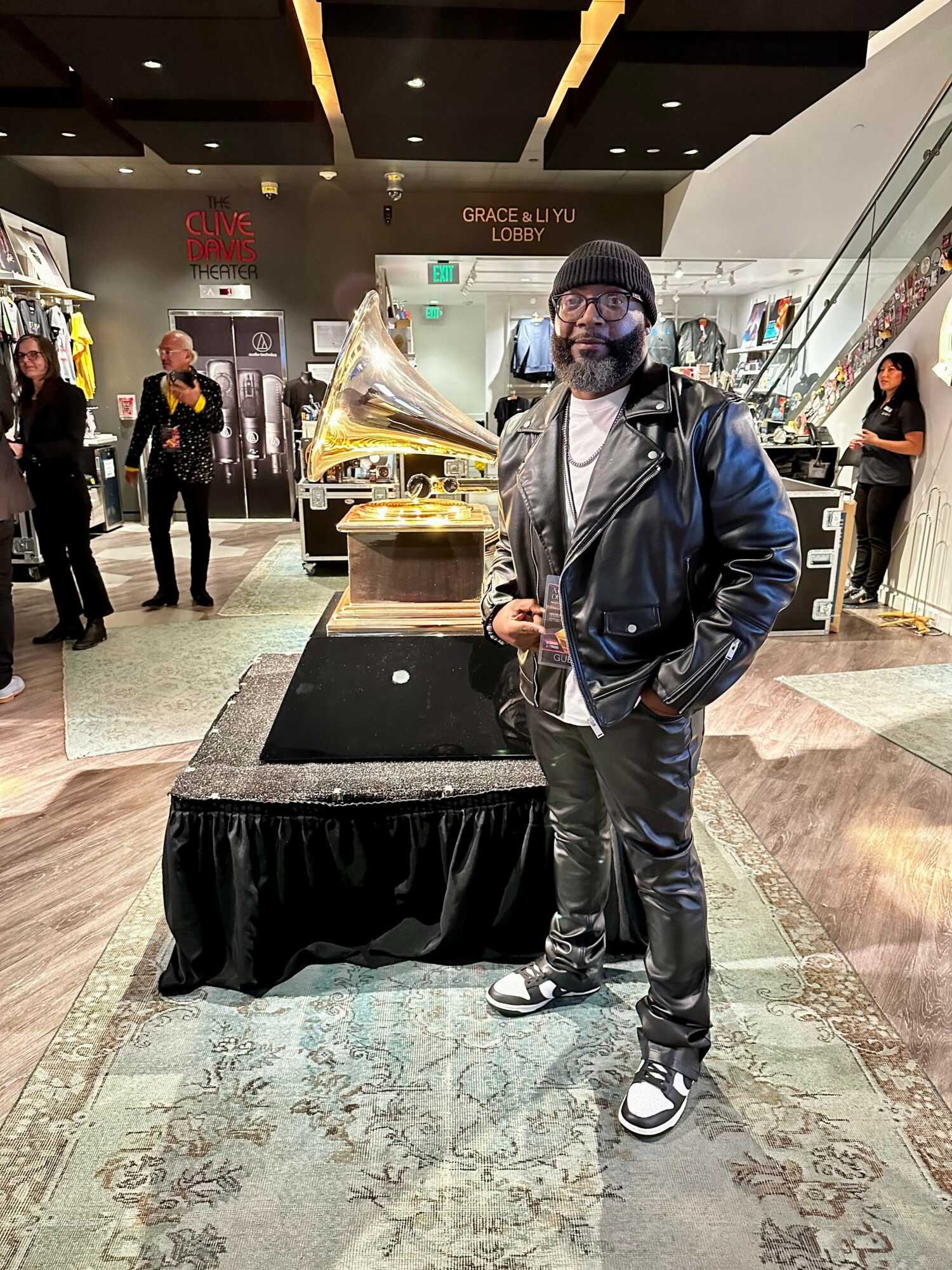 Man in black leather jacket and beanie stands in a shopping mall near a vintage gramophone display, with stores and people in background.