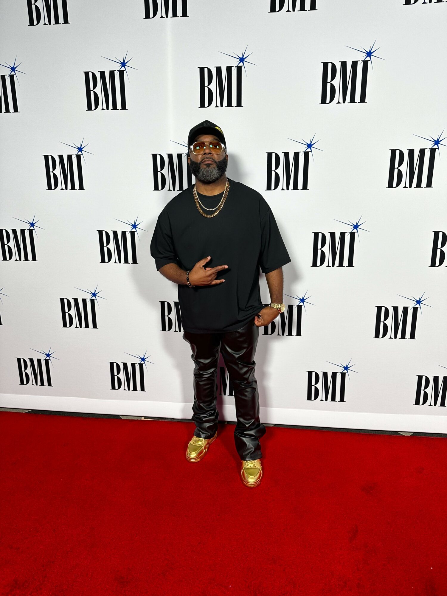 Person standing on red carpet in front of a BLI backdrop, wearing black top, patterned pants, and gold shoes.