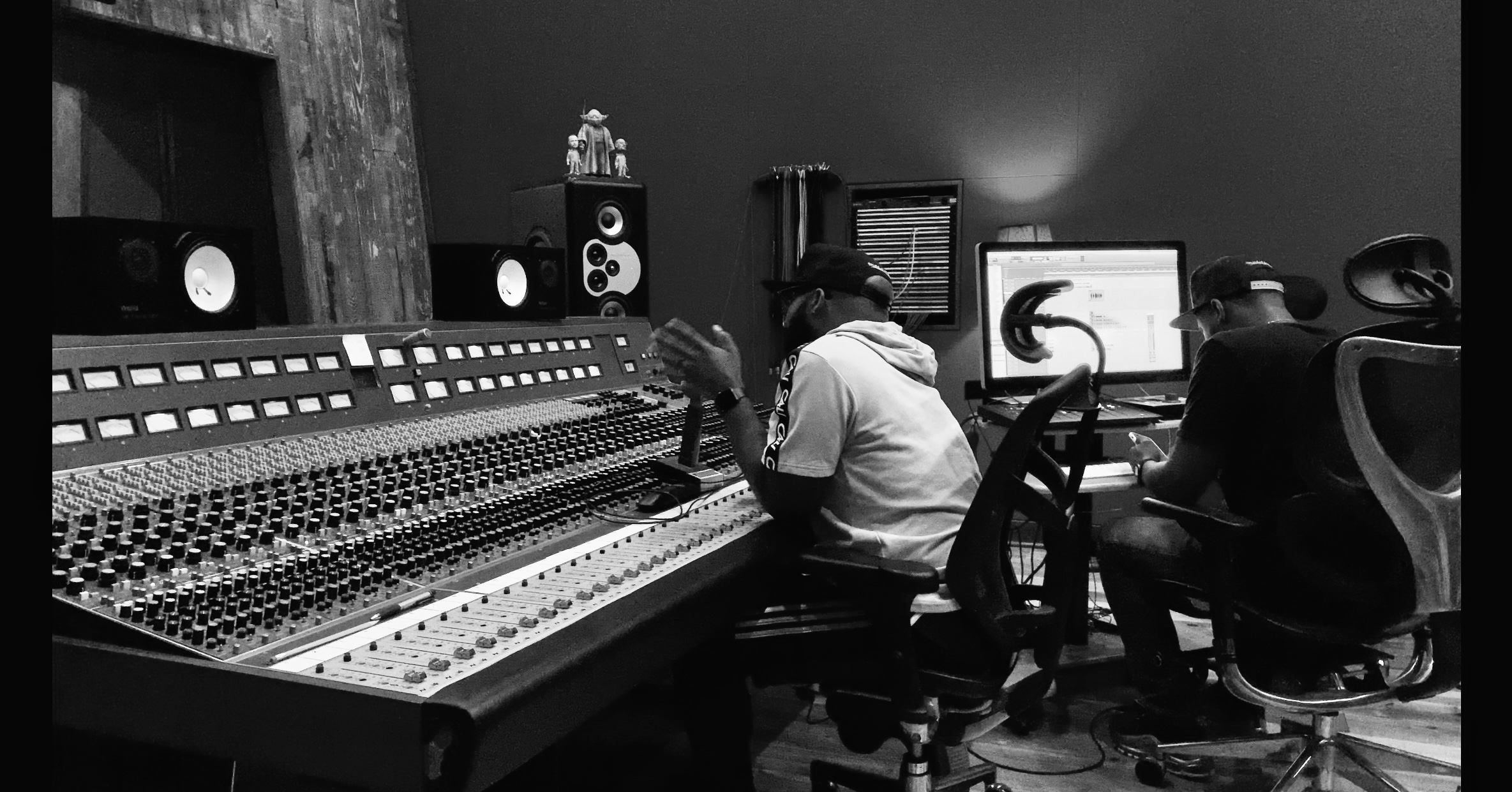 Two people working at a large audio mixing console in a recording studio, with computer monitors and speakers.
