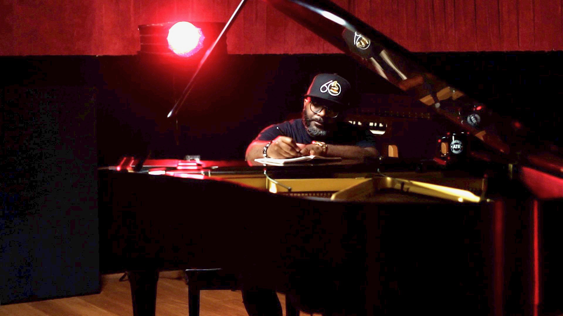 Person playing piano in a dimly lit room with red lighting, wearing a cap and glasses.