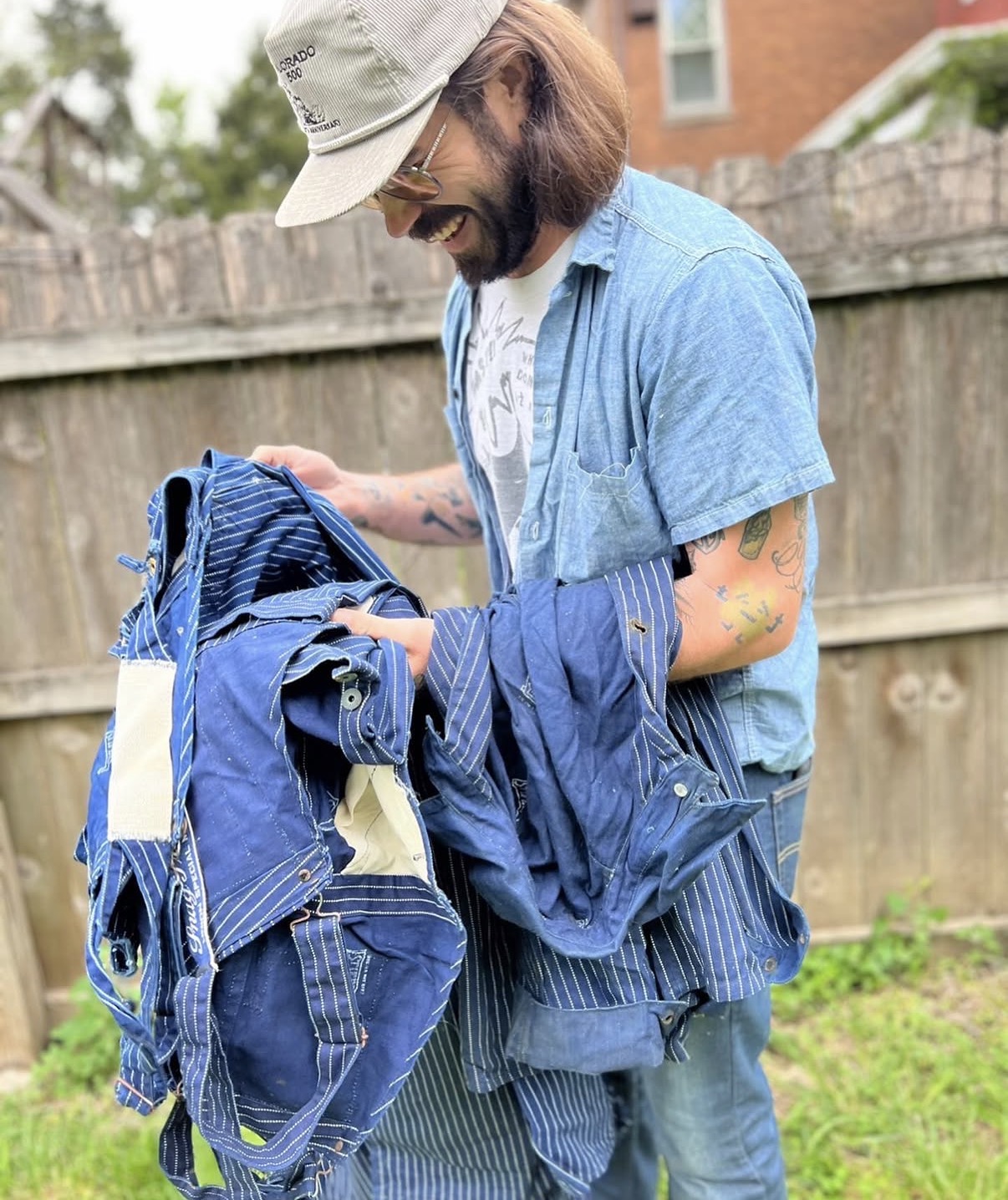 Person with a hat and beard holding blue clothing outdoors, smiling, with a wooden fence and grass in background.