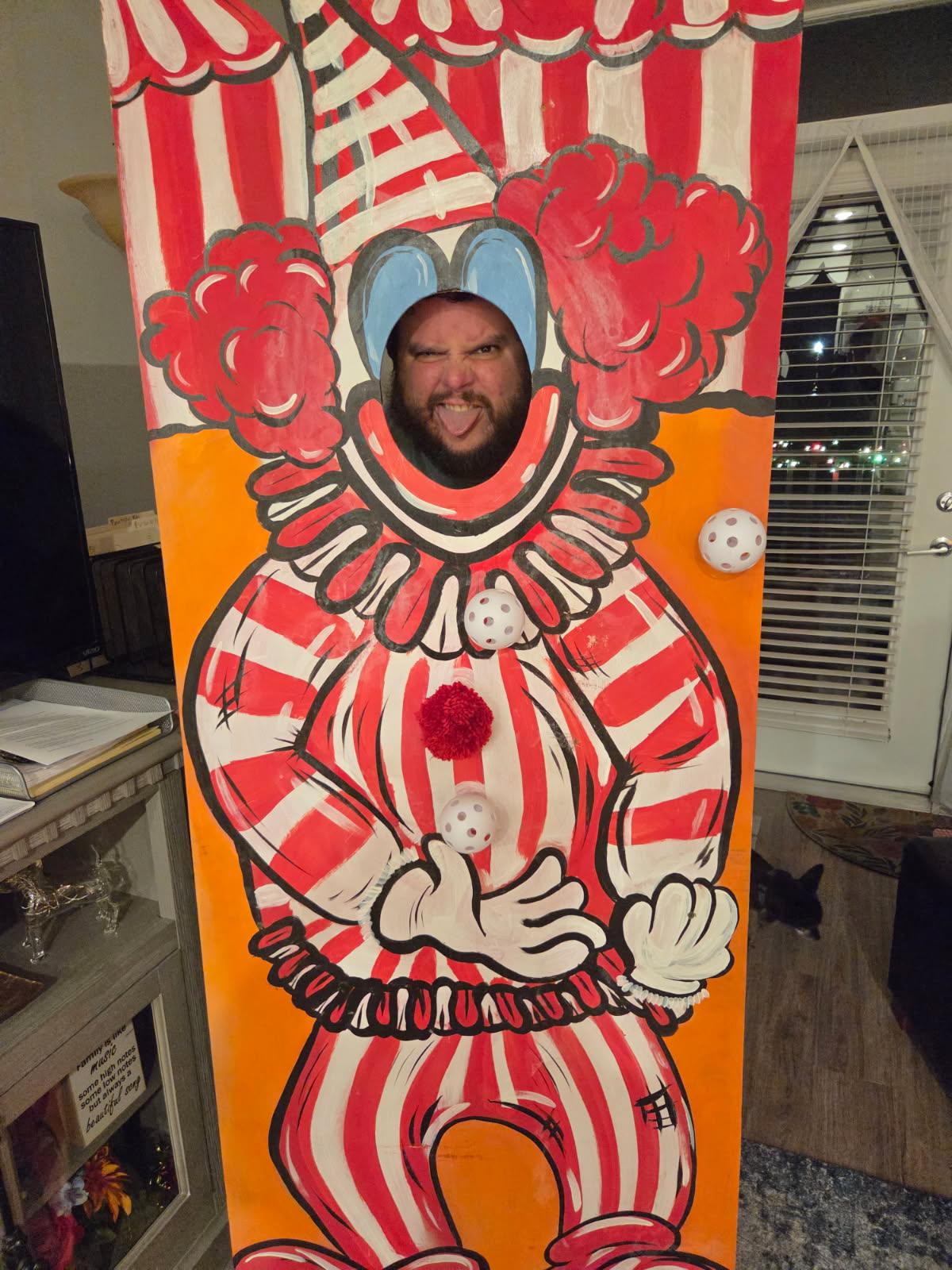 Person's face through a clown cutout with red and white striped costume, red hair, and a big red nose, smiling.