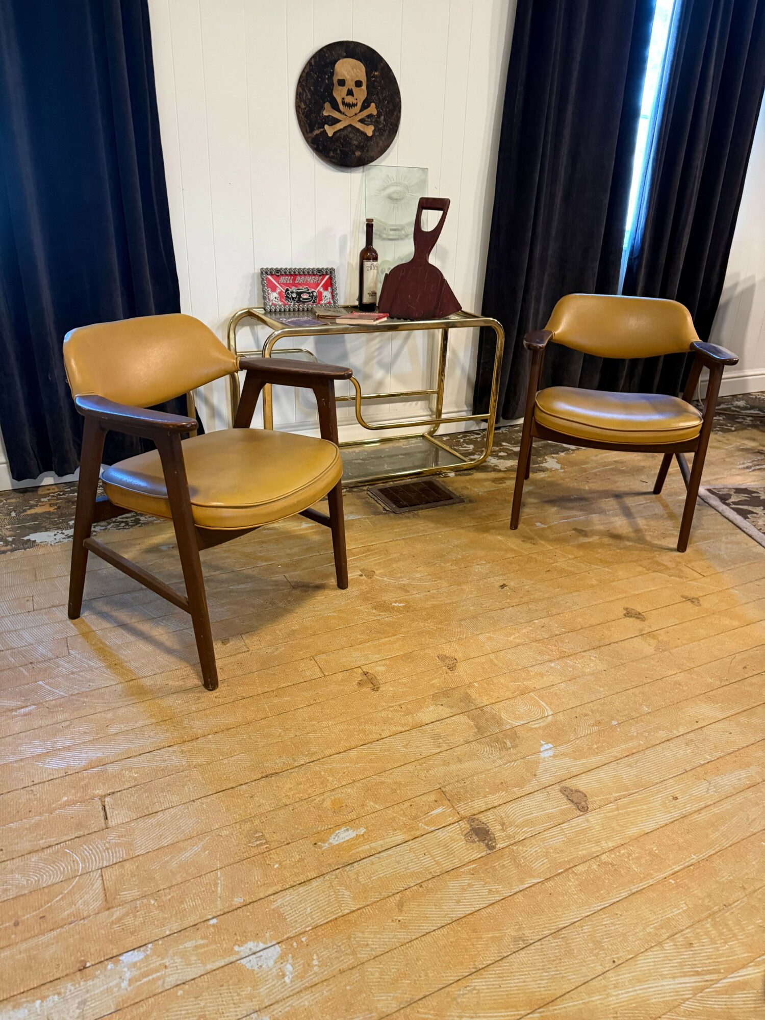 Three chairs with yellow seats and wooden legs are arranged in a room with wooden flooring and dark curtains. A wall decoration and a small table are visible.