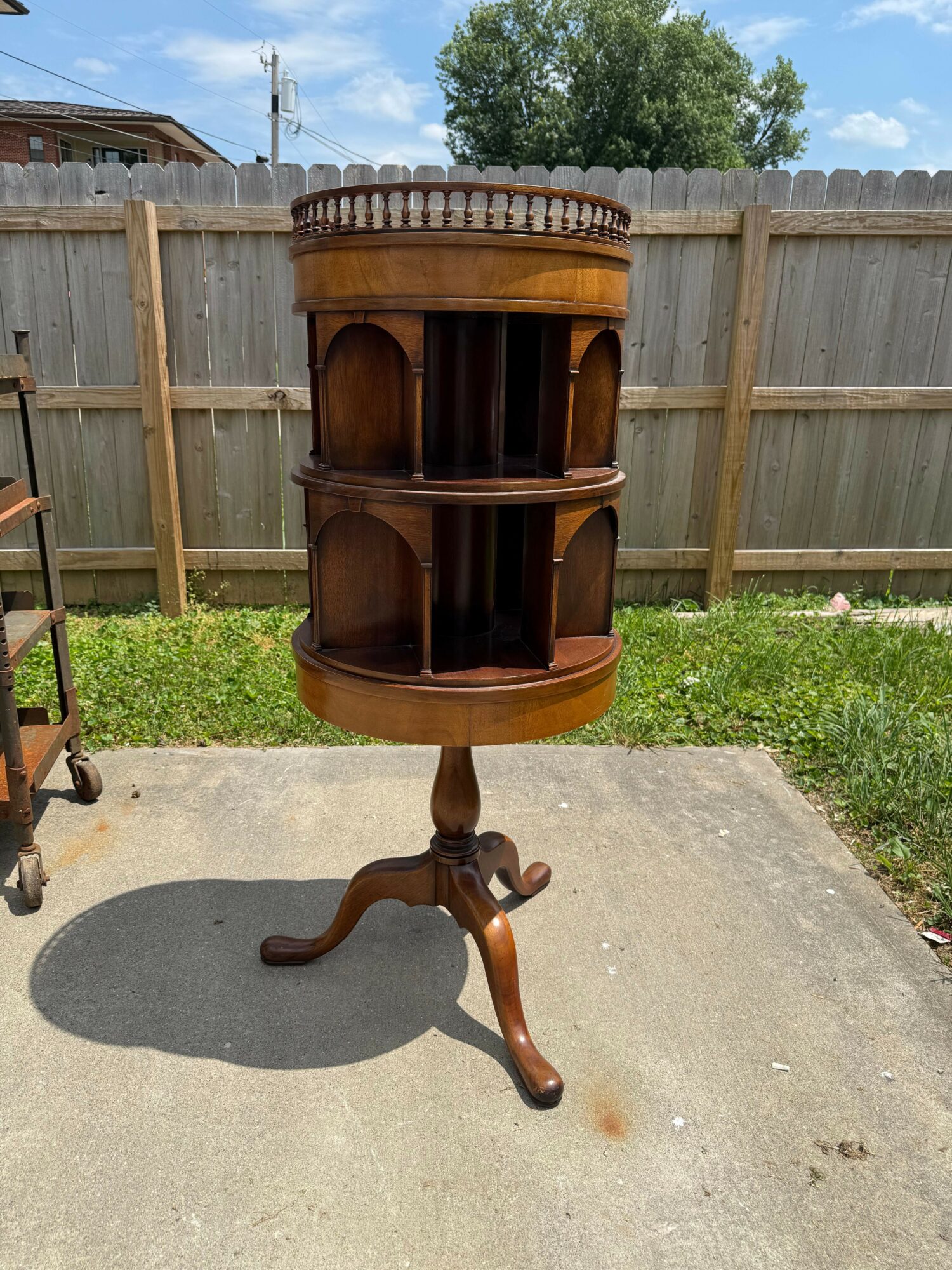 Round wooden bar cart with shelves and a curved leg, placed outdoors on concrete with a wooden fence and trees in background.