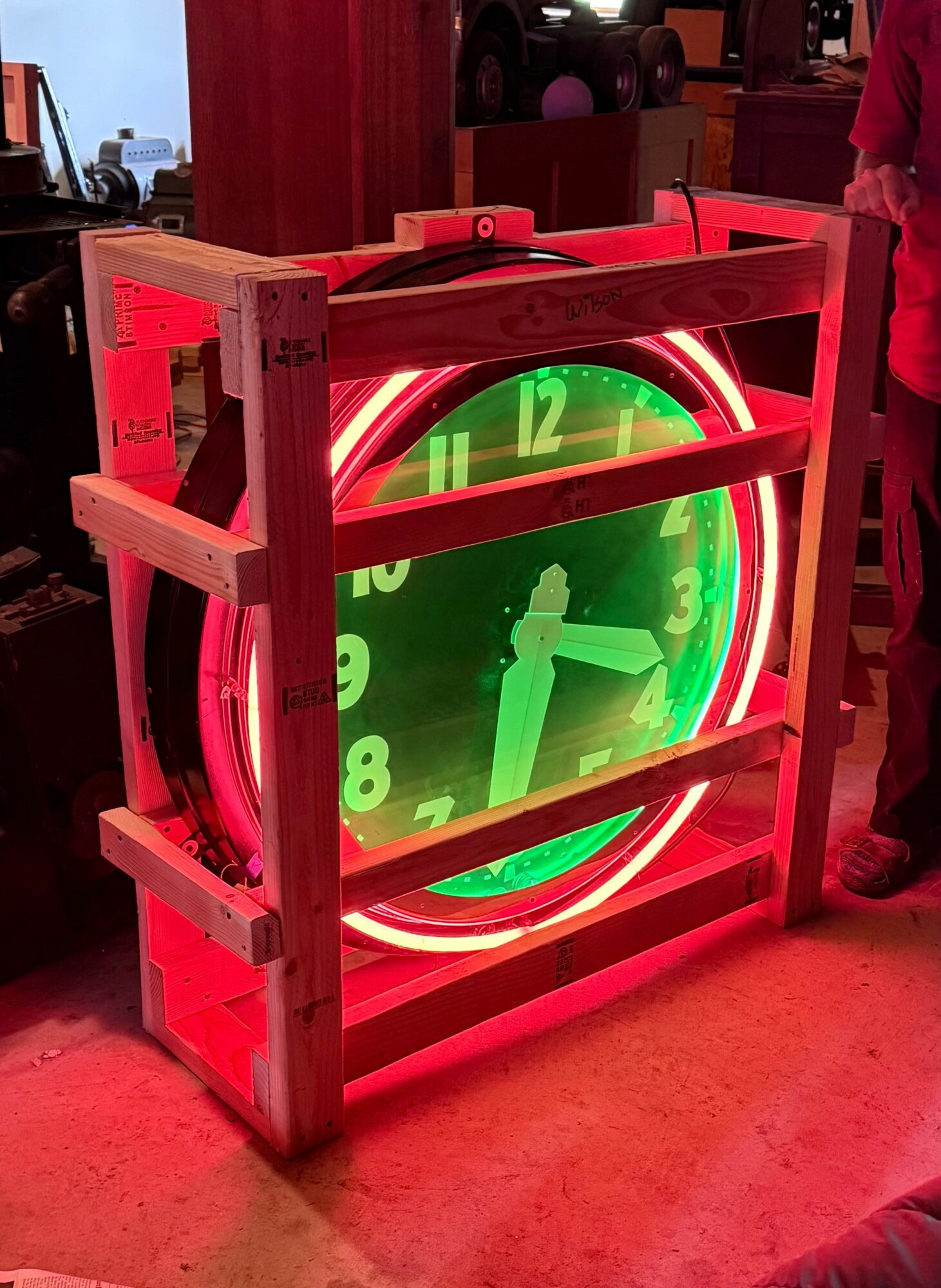 Neon clock with green face and white hands inside a wooden frame, illuminated with red light, in a room setting.
