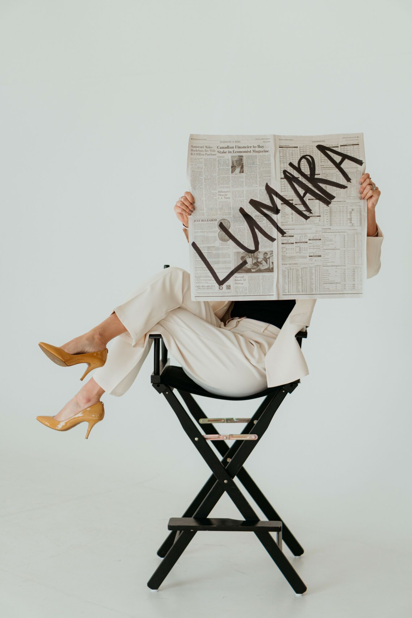 Person sitting on a black chair, holding a newspaper with 'LUMAR' written on it, legs crossed, against a plain background.