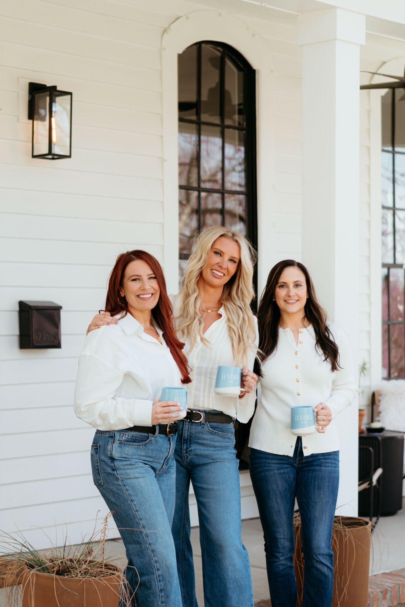 Three women stand outside a white building holding drinks, smiling, with large windows and a lantern on the wall.