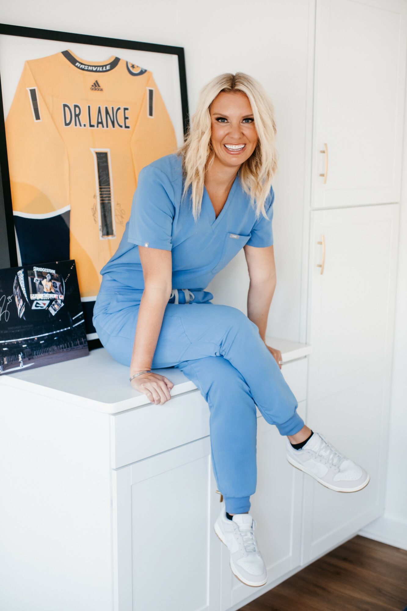 Smiling woman in blue scrubs sitting on a white cabinet, with a framed sports jersey behind her.