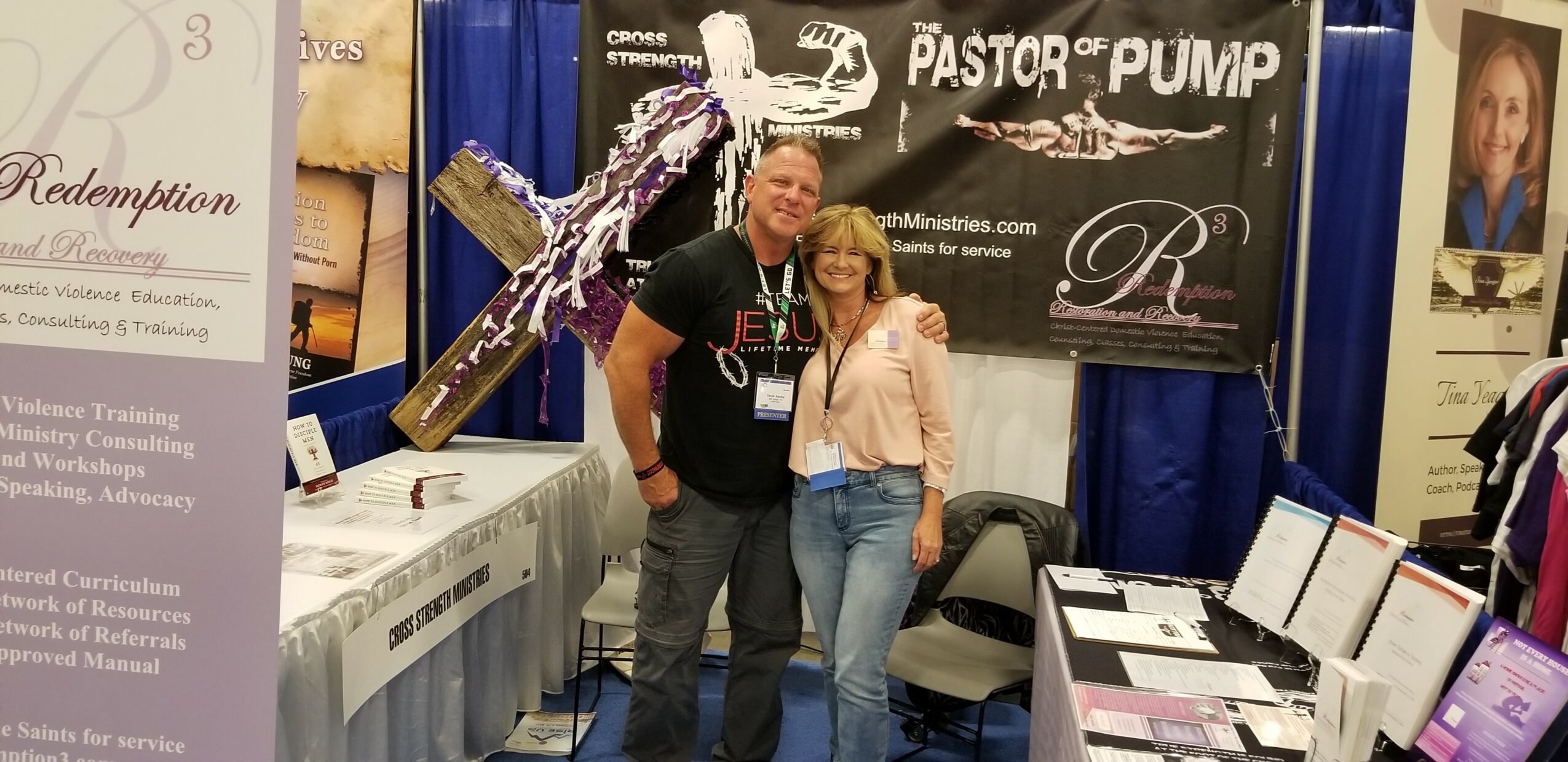 Two people standing together at a booth, smiling, with banners and tables in the background.