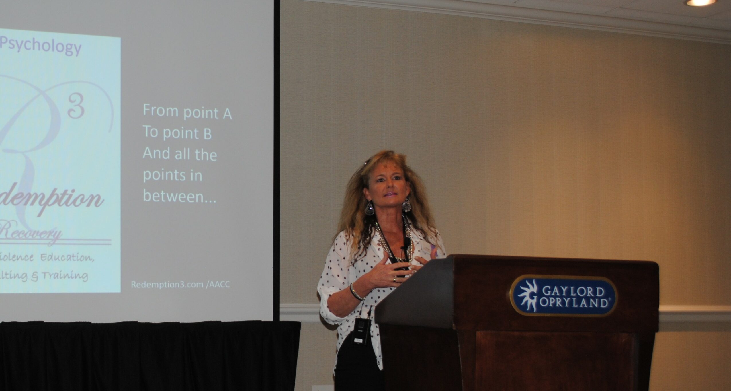Woman speaking at a podium in a conference room with a presentation slide projected behind her.