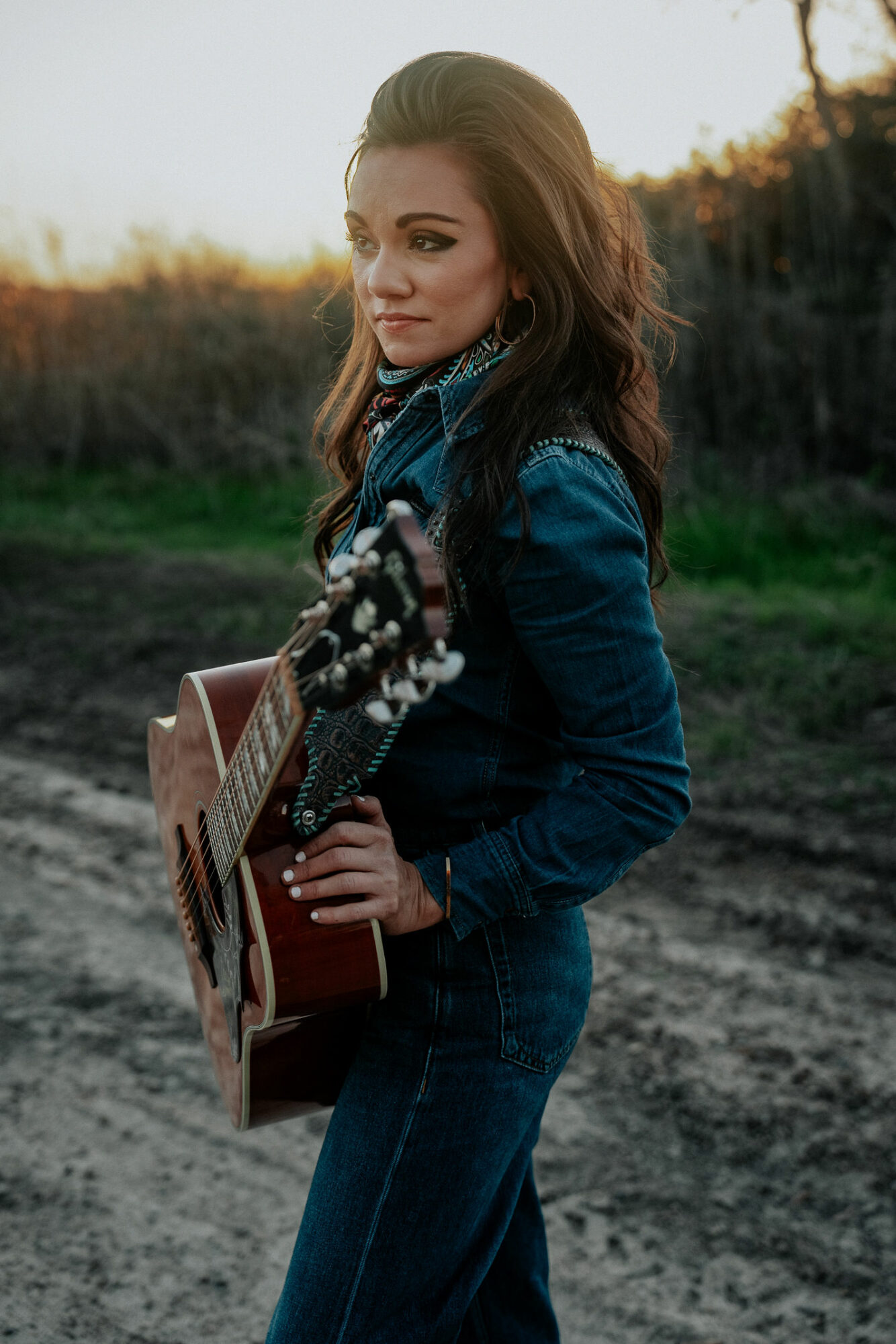 Young woman with long hair holding an acoustic guitar outdoors during sunset, standing on a dirt path.
