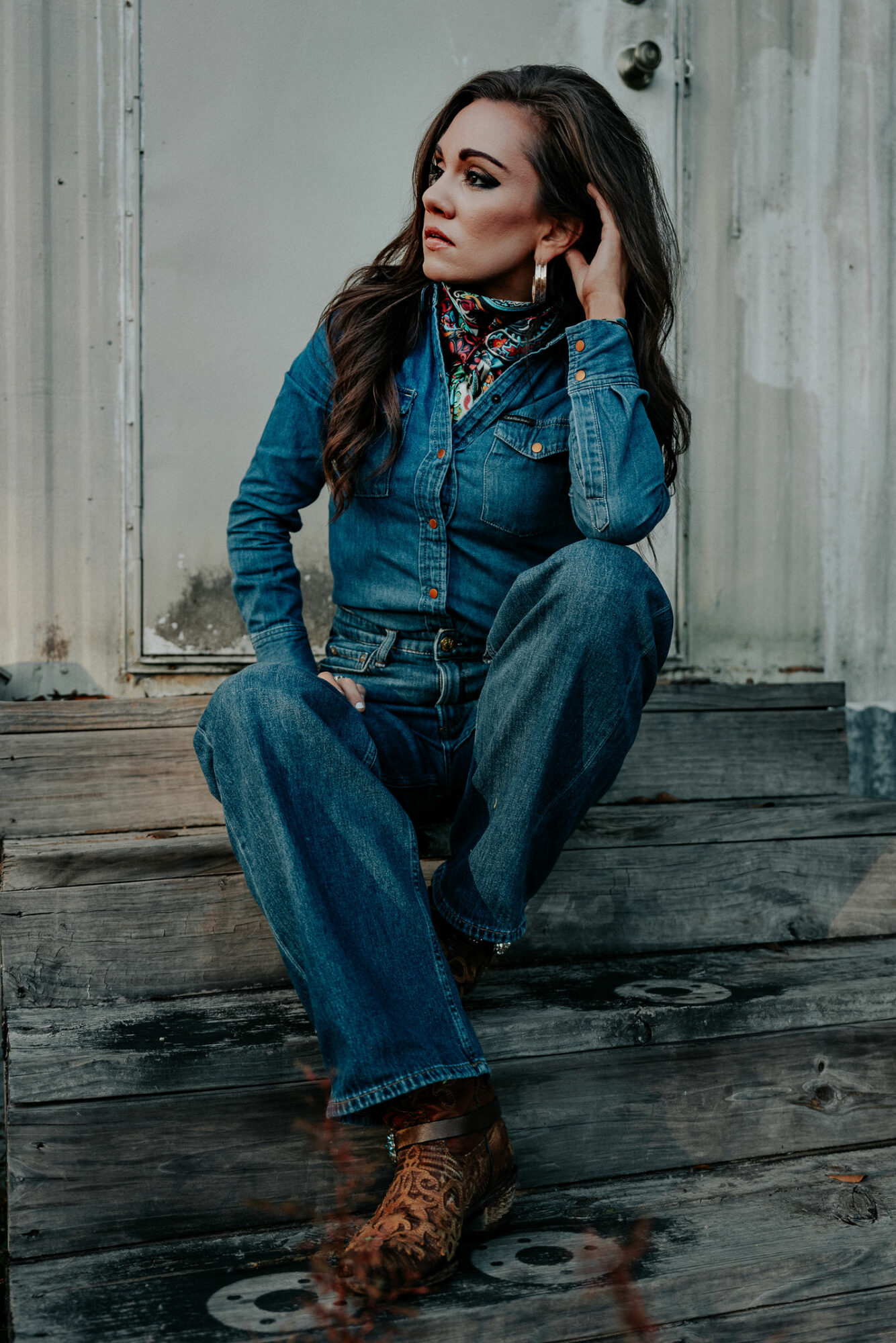 Woman sitting on wooden steps, wearing denim outfit and patterned boots, looking to the side, with a neutral background.